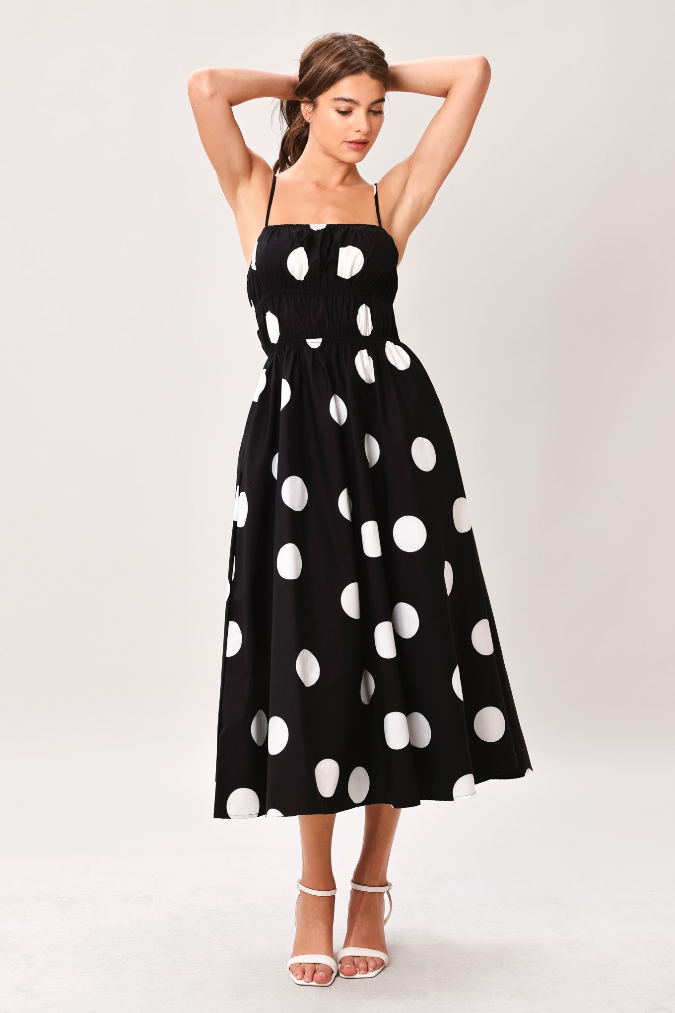 Woman wearing a black midi dress with white polka dots featuring straight neckline, straps, elastic at empire and waist, side pockets and full skirt | Flying Tomato