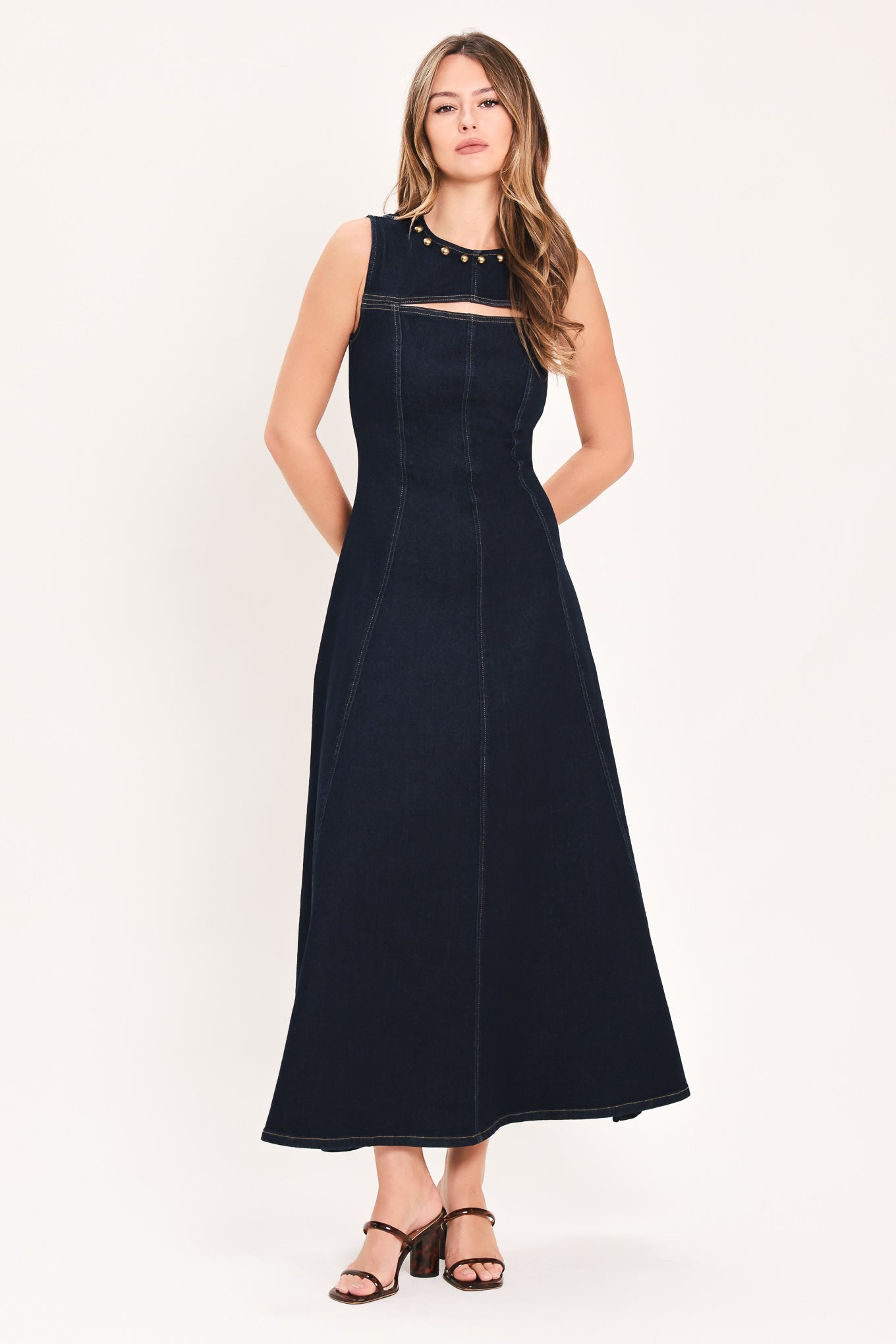 A woman wearing a sleeveless dark navy maxi dress with a crew neckline, a horizontal cutout across the chest, and small gold stud embellishments | Flying Tomato