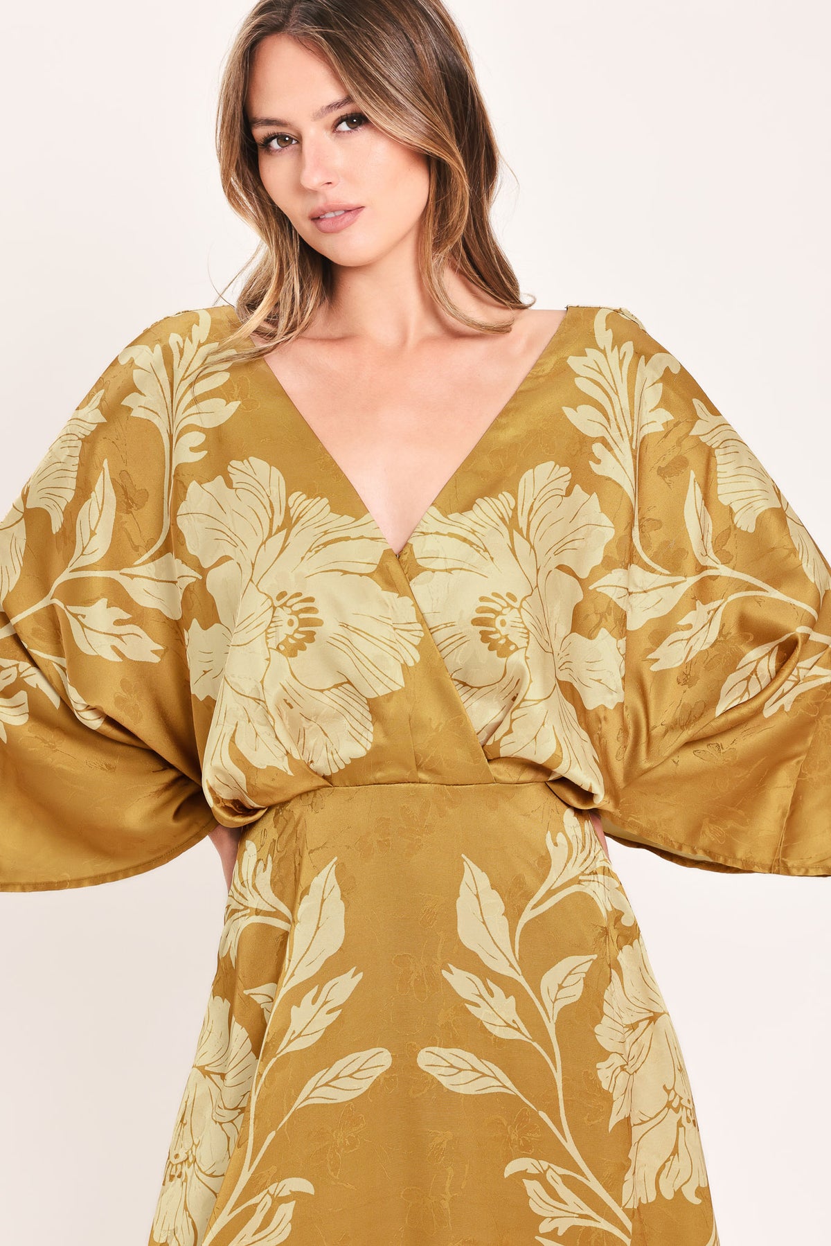 A woman wearing a midi-length, V-neck dress with wide bell sleeves in a golden camel fabric featuring a cream floral jacquard pattern | Flying Tomato