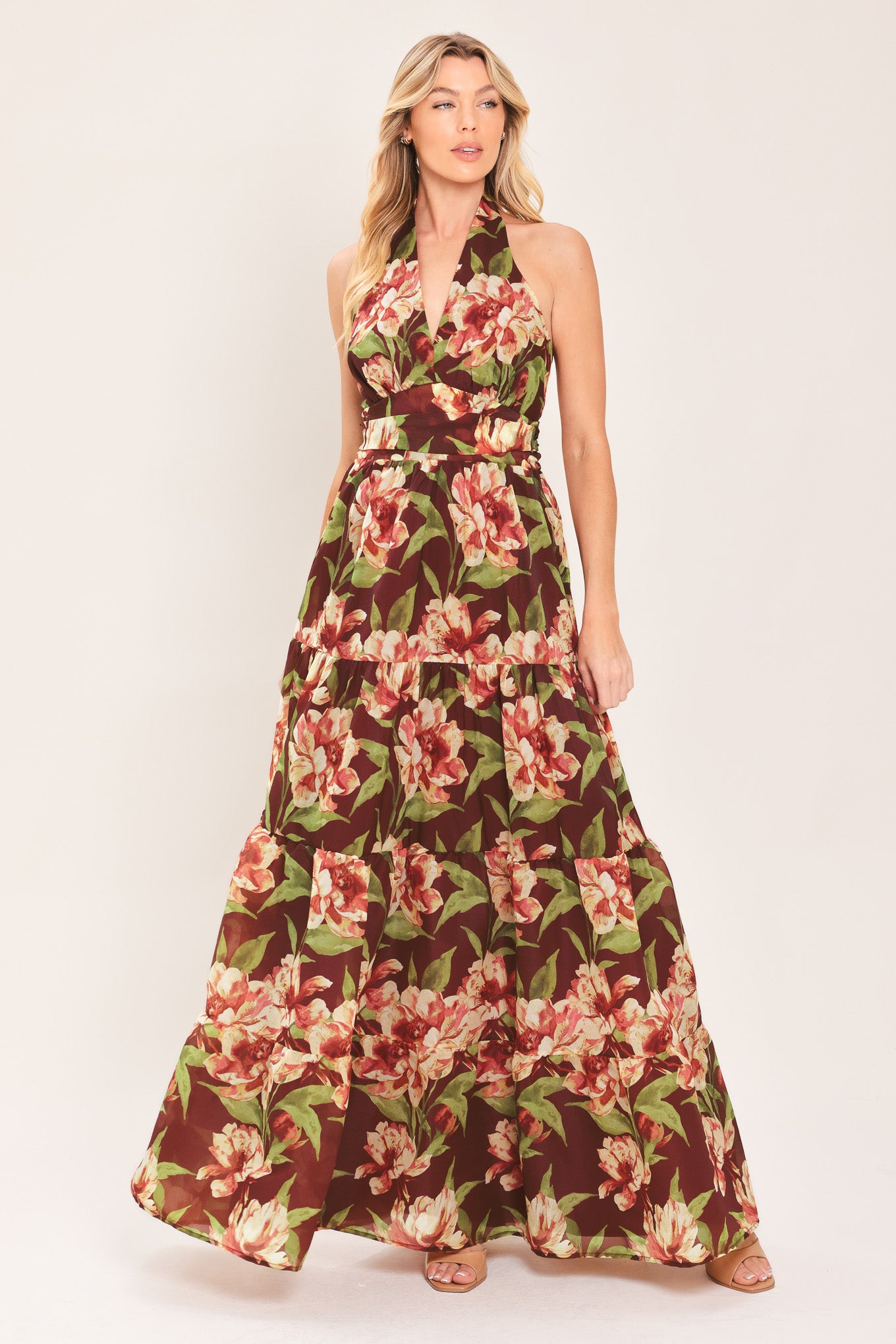 A woman wearing a halter-neck, tiered maxi dress with a burgundy base and a large pink and green floral print | Flying Tomato