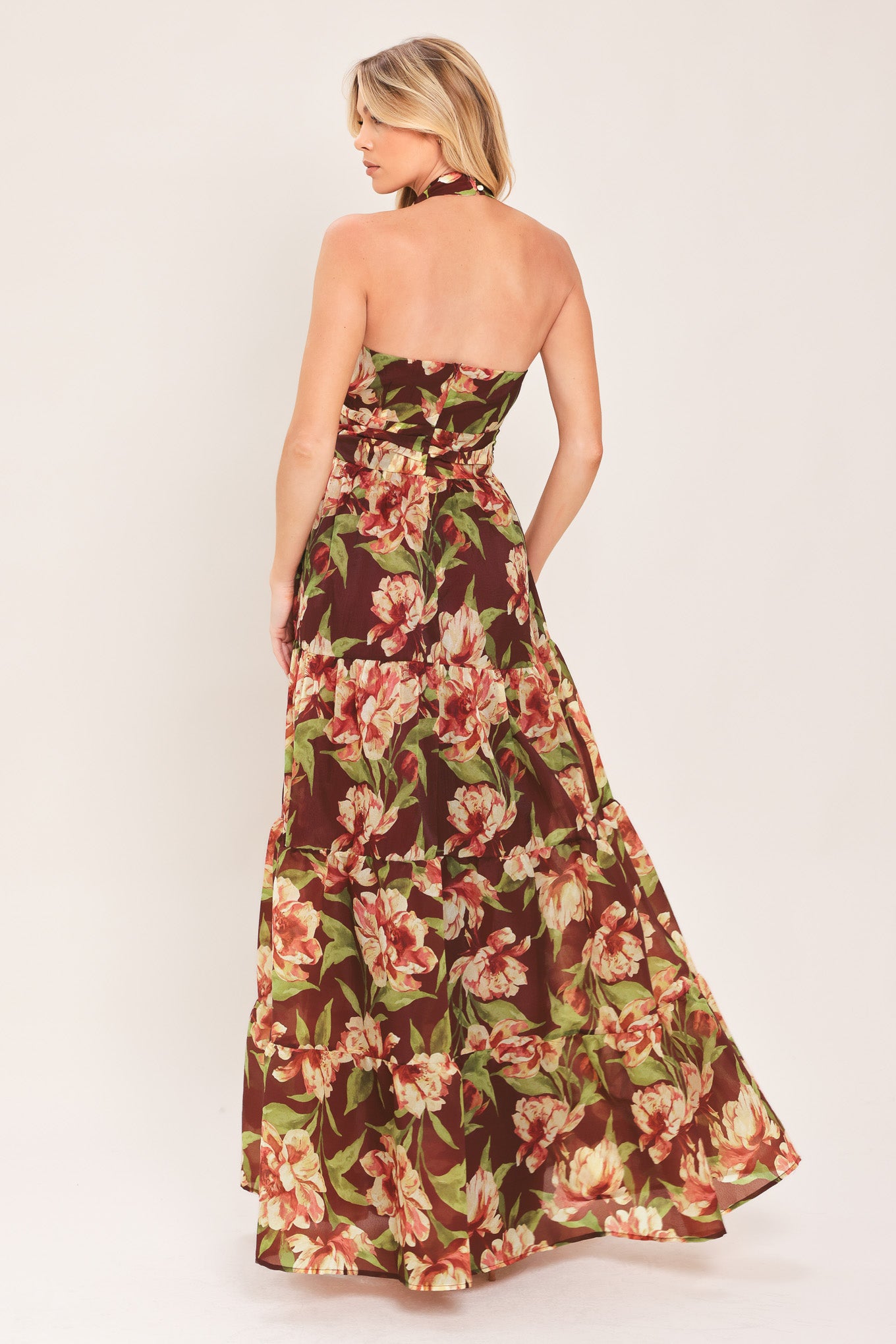 A woman wearing a halter-neck, tiered maxi dress with a burgundy base and a large pink and green floral print | Flying Tomato