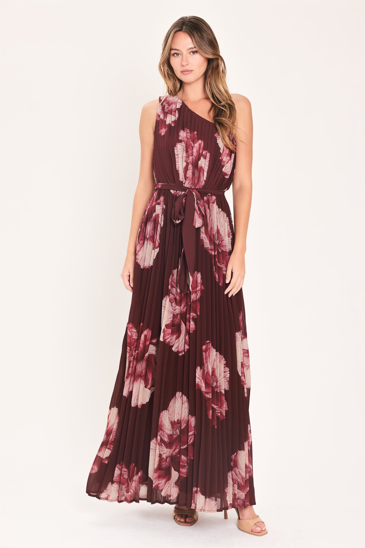A woman modeling a sleeveless, one-shoulder maxi dress with a pleated skirt and waist tie. The dress features a dark burgundy base with a large floral print in dusty pink and mauve tones | Flying Tomato