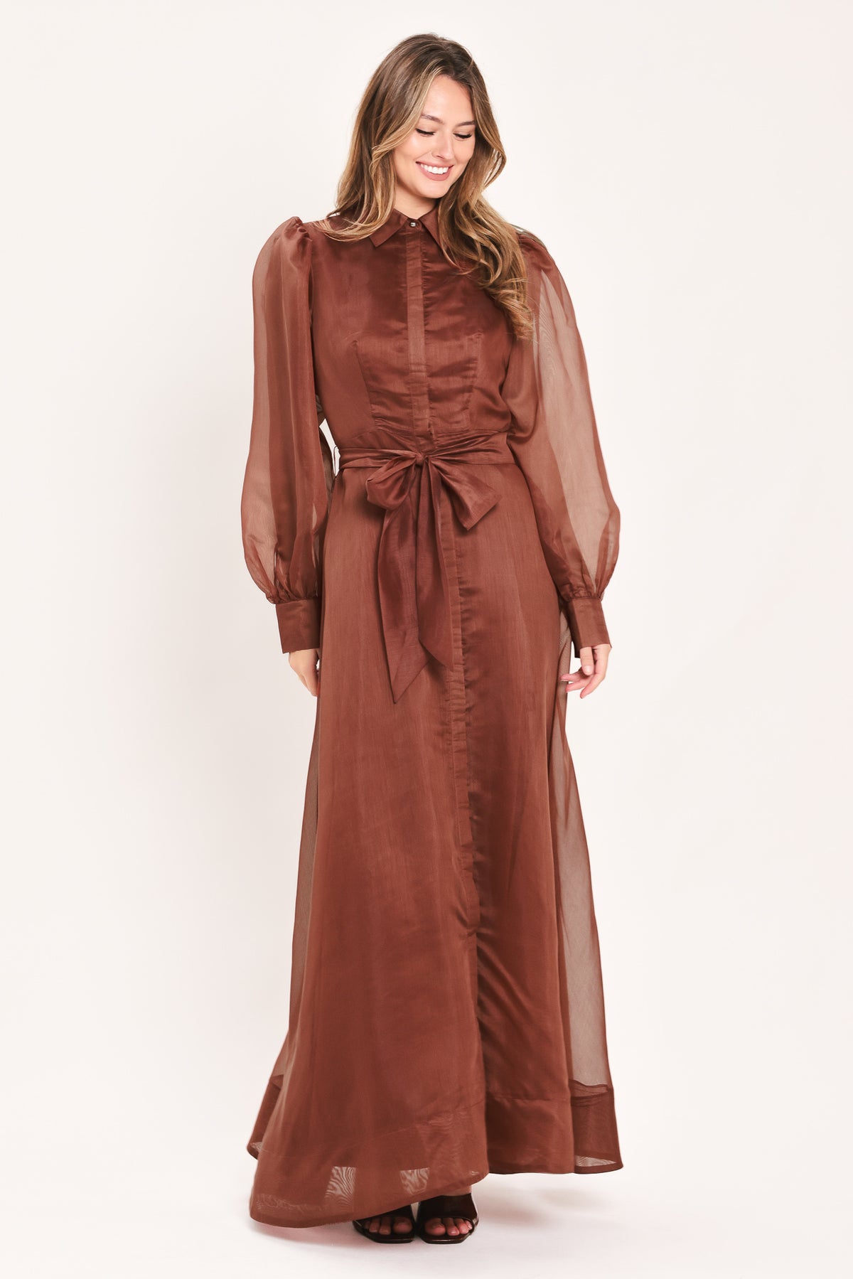 A woman wearing a rust-brown maxi dress with sheer balloon sleeves, a high ruffled collar, and a matching fabric tie belt at the waist | Flying Tomato