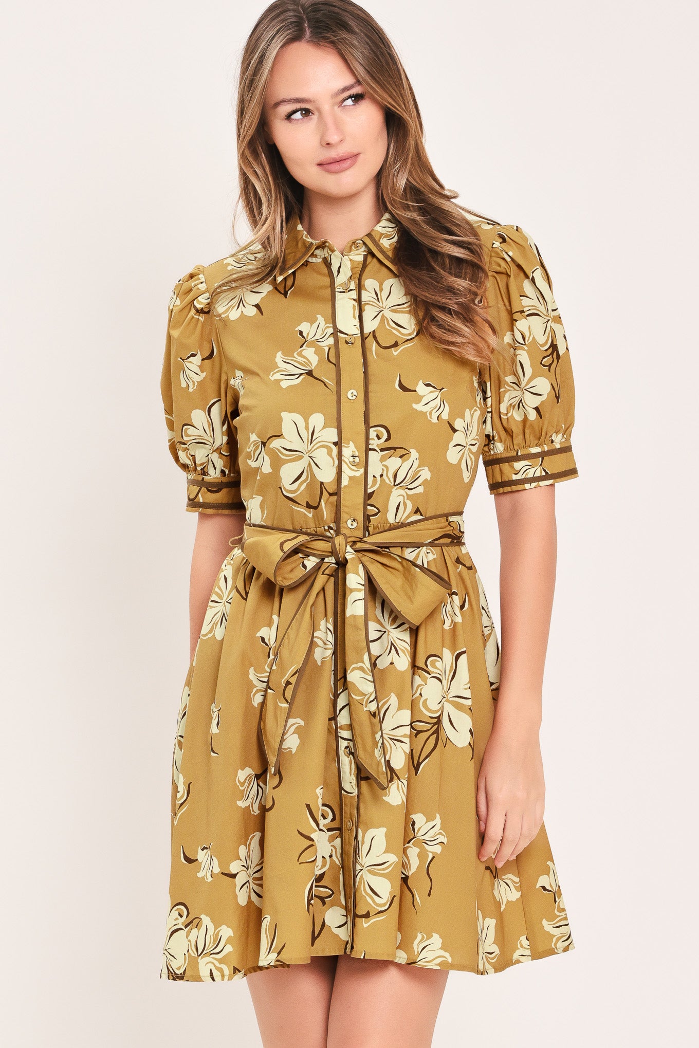 A woman wearing a camel yellow short-sleeve button-up dress with a beige and white floral print, featuring a tie waist belt | Flying Tomato
