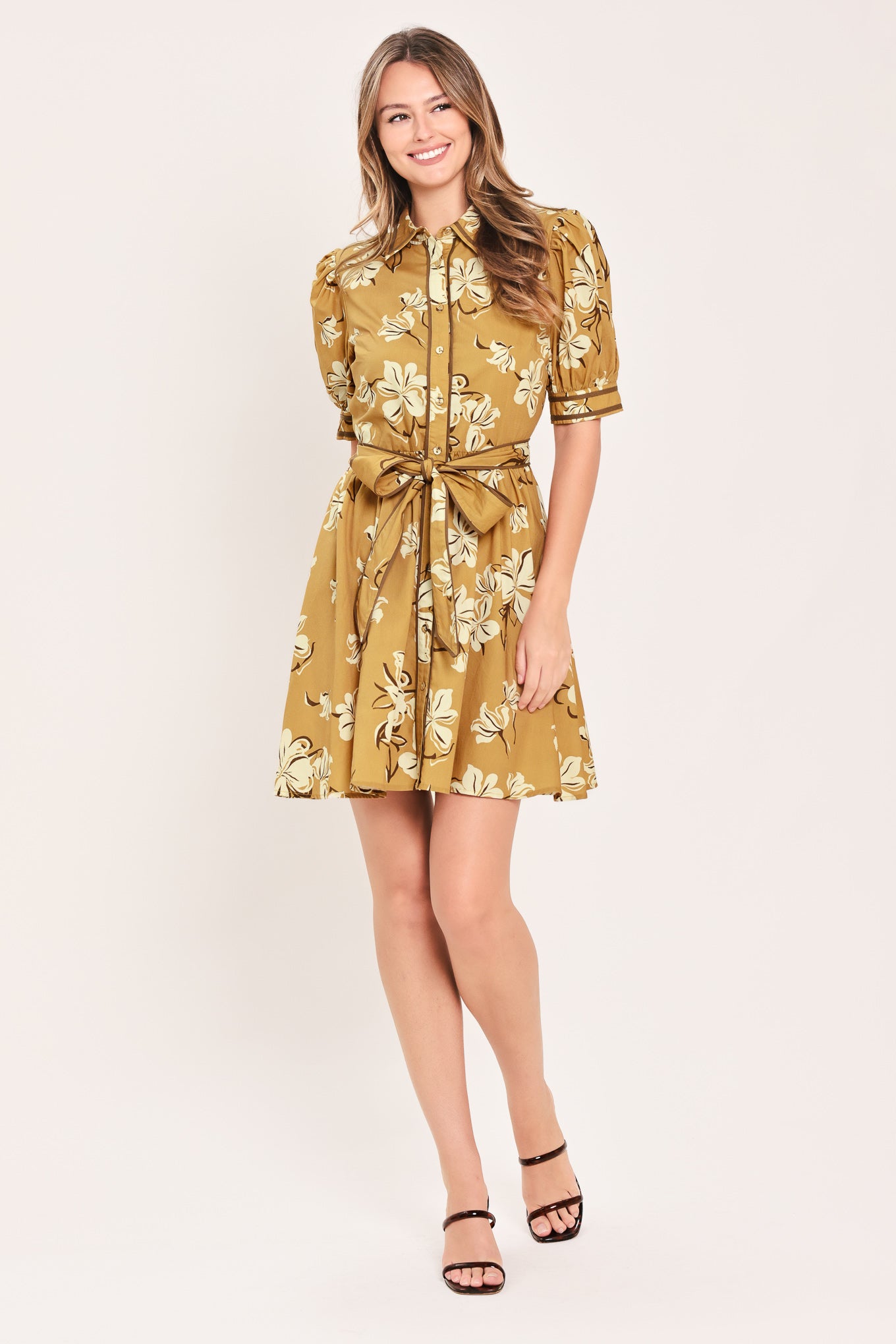 A woman wearing a camel yellow short-sleeve button-up dress with a beige and white floral print, featuring a tie waist belt | Flying Tomato