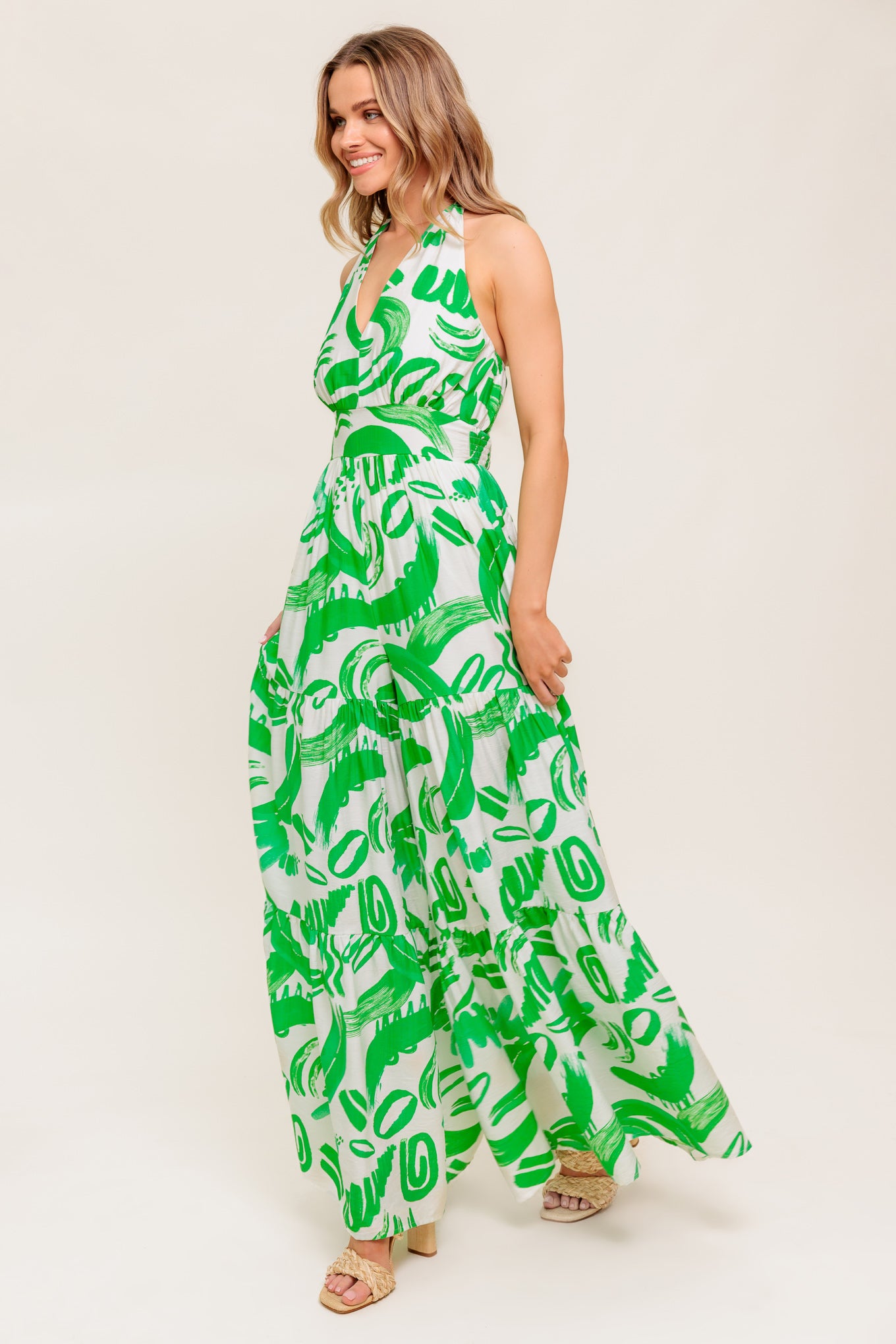 A printed woven jumpsuit featuring halter neckline, tiered pant, bare back with tie and smocked back waistband
