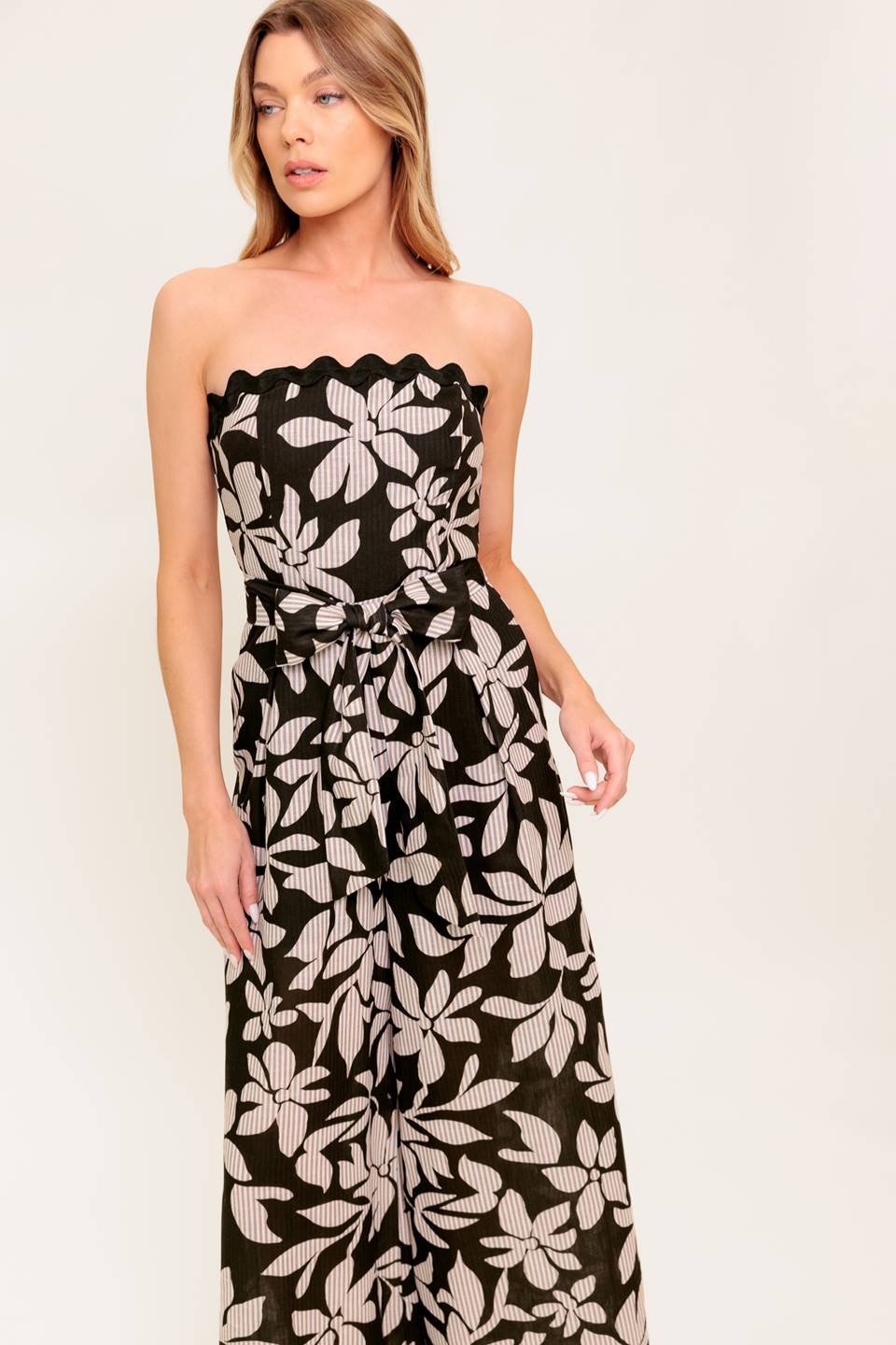 A printed woven jumpsuit featuring strapless neckline with ric rac trimmed edge, self sash tie, wide leg and back zipper closure.