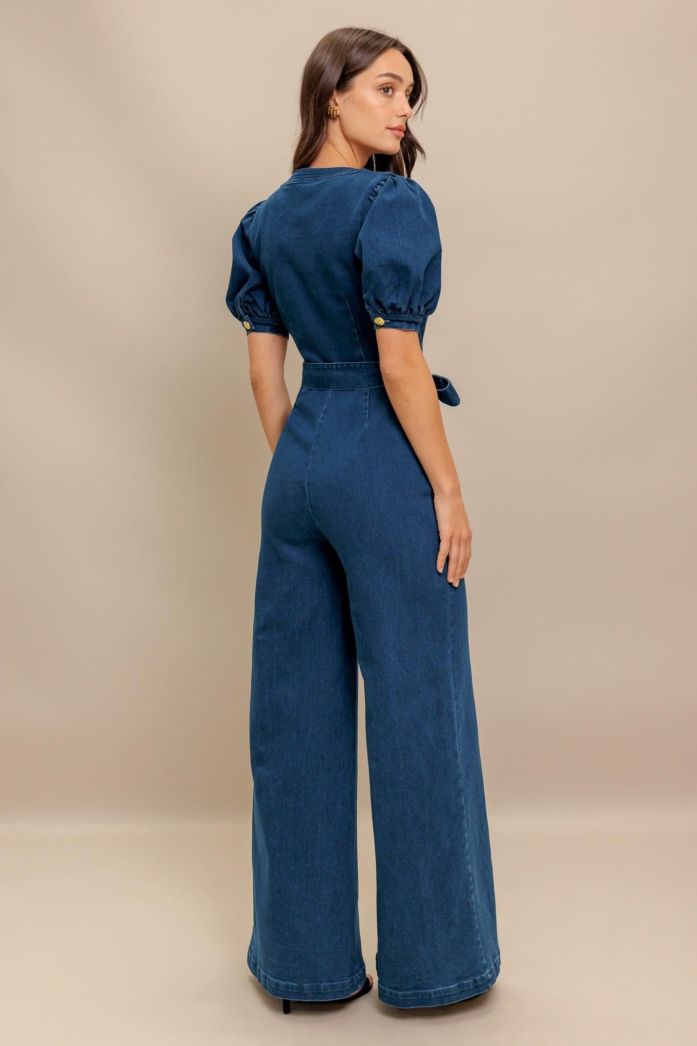 A washed denim jumpsuit featuring round neckline, short puff sleeve, front pockets, button down, self belt and wide leg