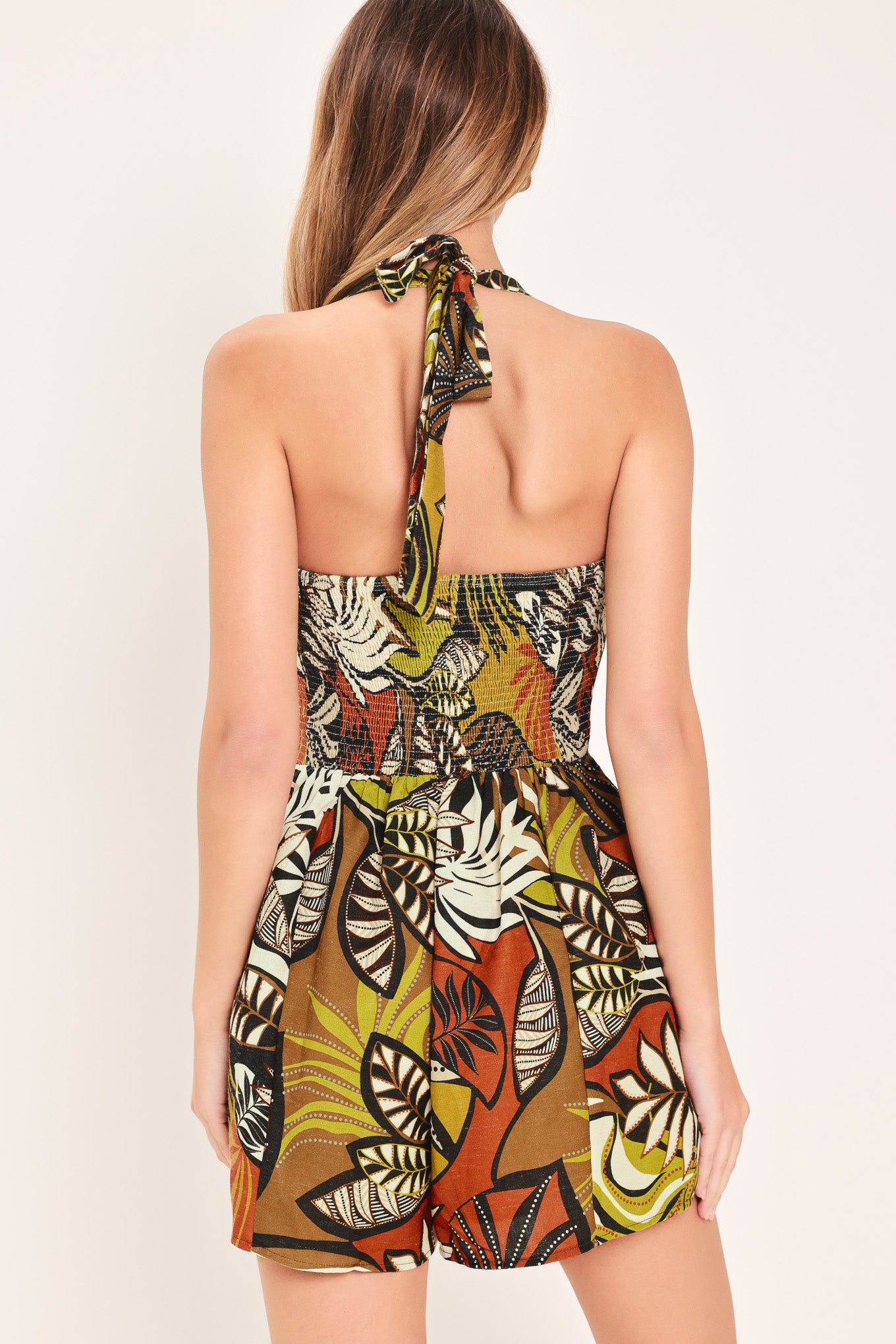Woman wearing a colorful brown and mustard yellow palm leaf patterned romper featuring halter neckline and smocked back bodice | Flying Tomato