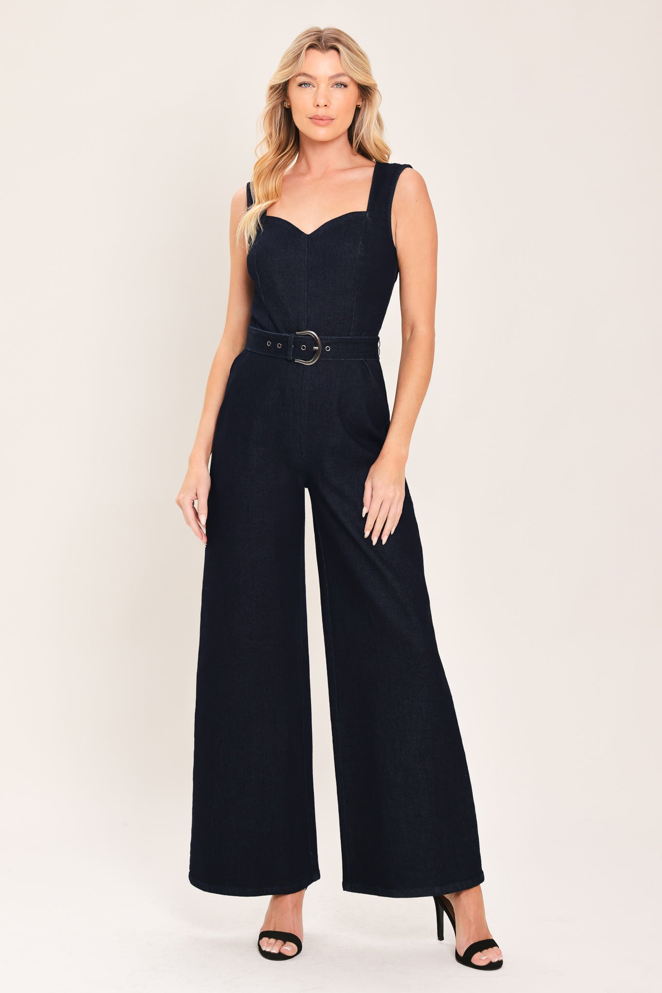 Woman wearing a dark indigo denim jumpsuit featuring sweetheart neckline, sleeveless, self belt, wide leg and back zipper closure | Flying Tomato