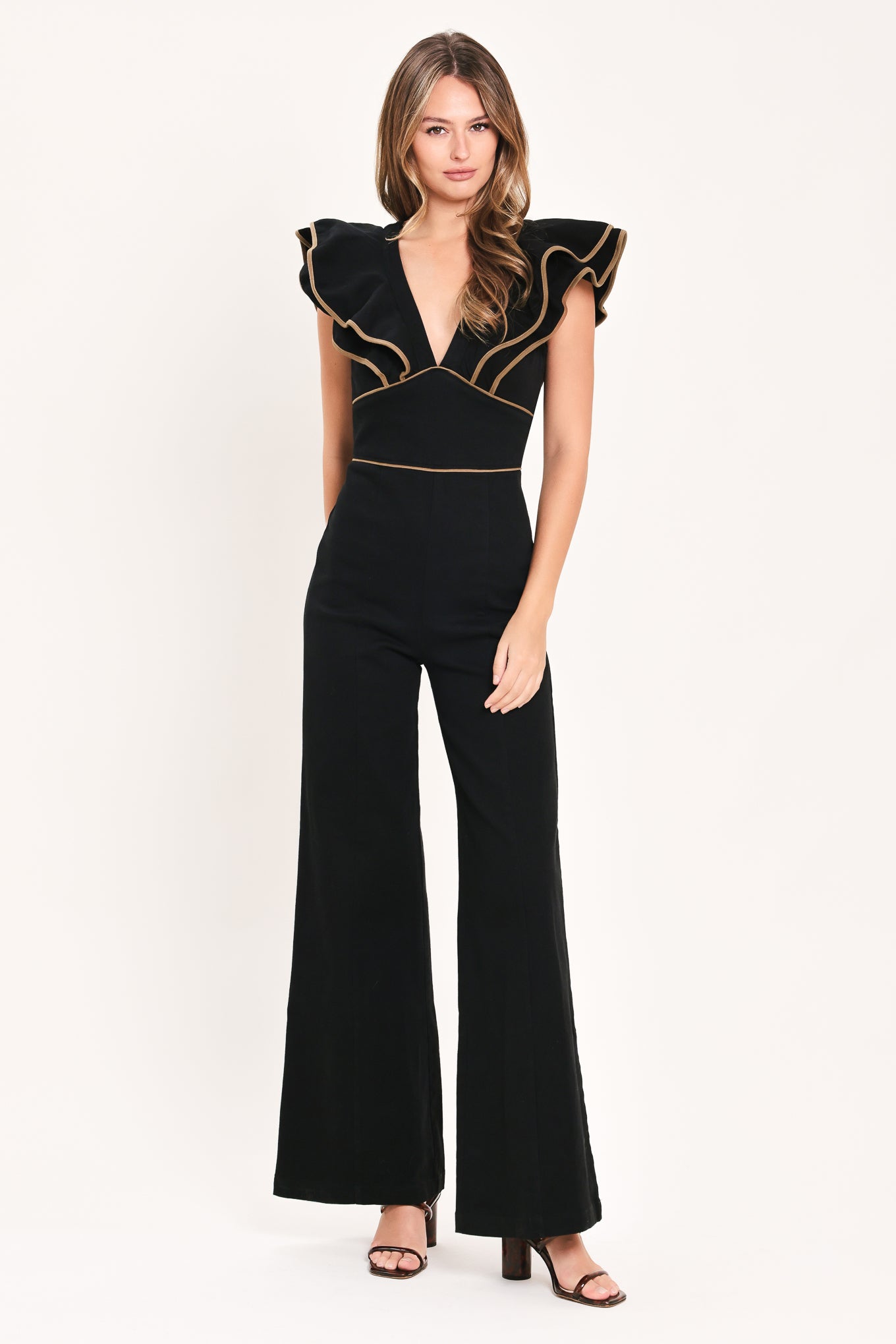 A woman modeling a black jumpsuit with dramatic ruffled cap sleeves, a deep V-neckline, and beige contrast piping details | Flying Tomato