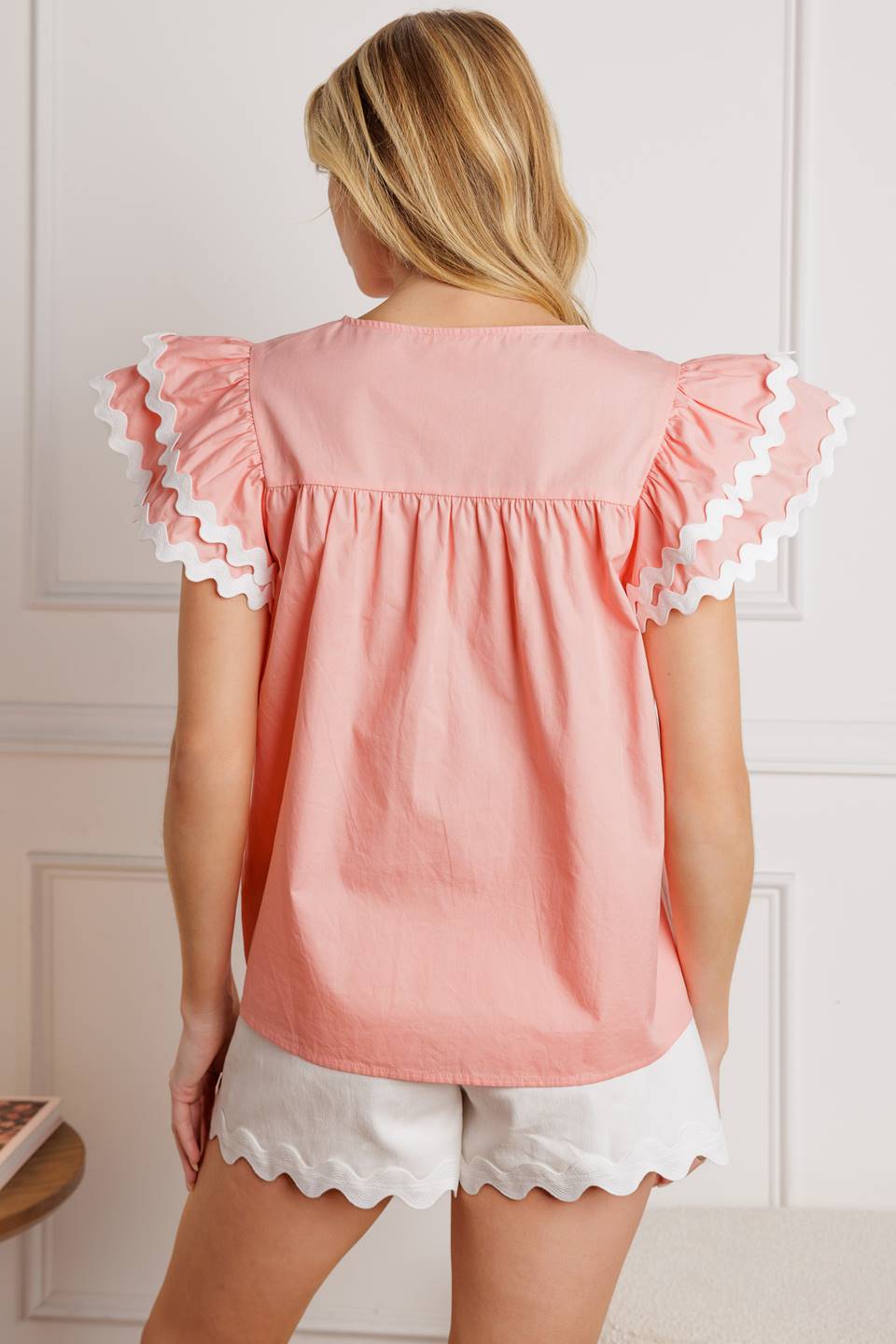 A woven top with a round neckline, tie and tassel, and double ruffled sleeves with ric rac trimming.
