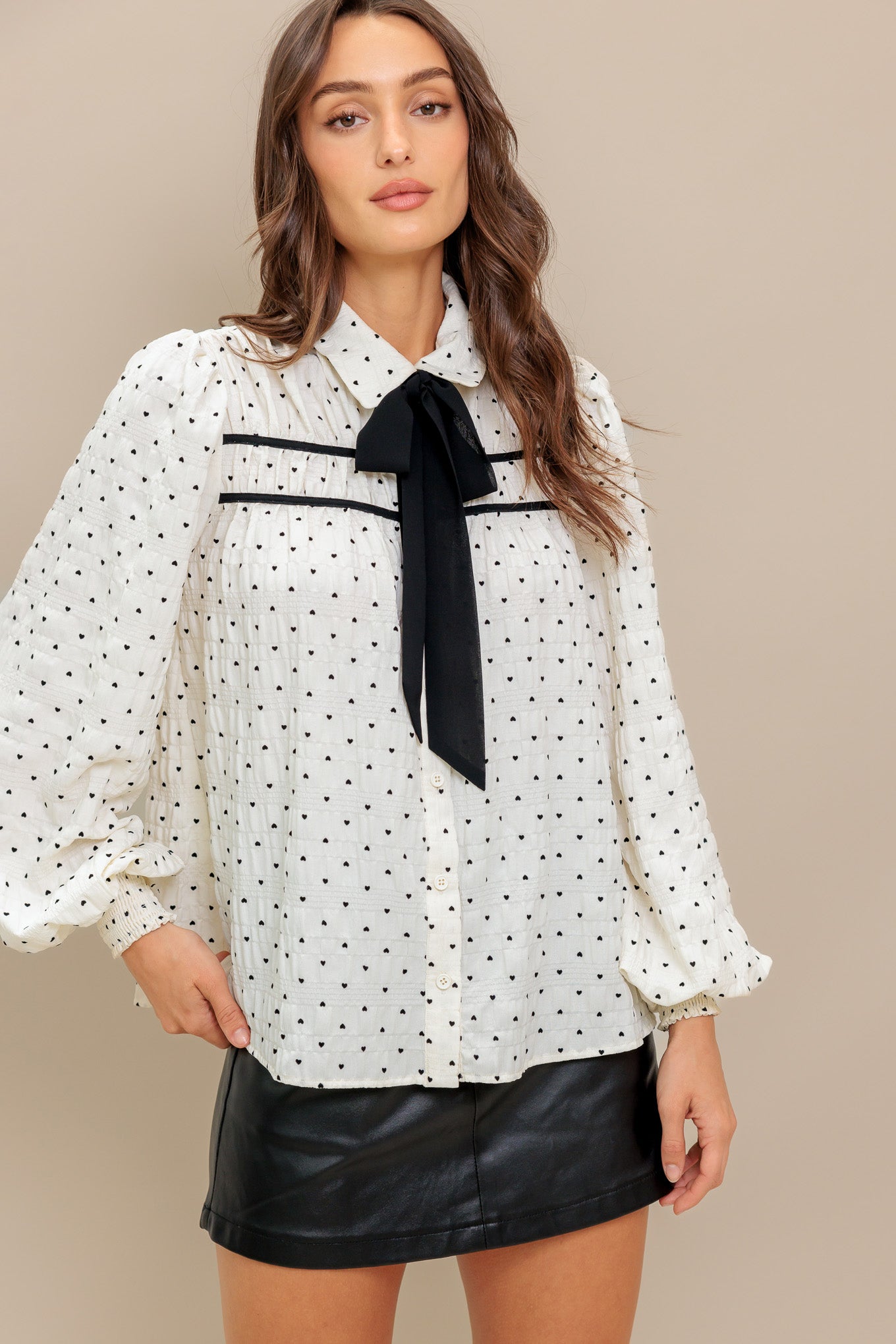 A heart printed woven top featuring collar, contrasting bow tie, long sleeve with smocked cuff and front button closure