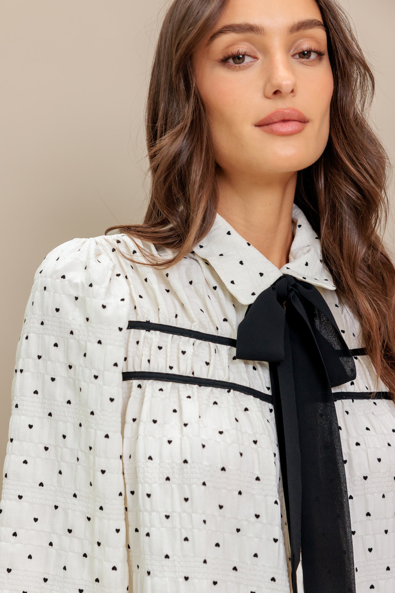 A heart printed woven top featuring collar, contrasting bow tie, long sleeve with smocked cuff and front button closure
