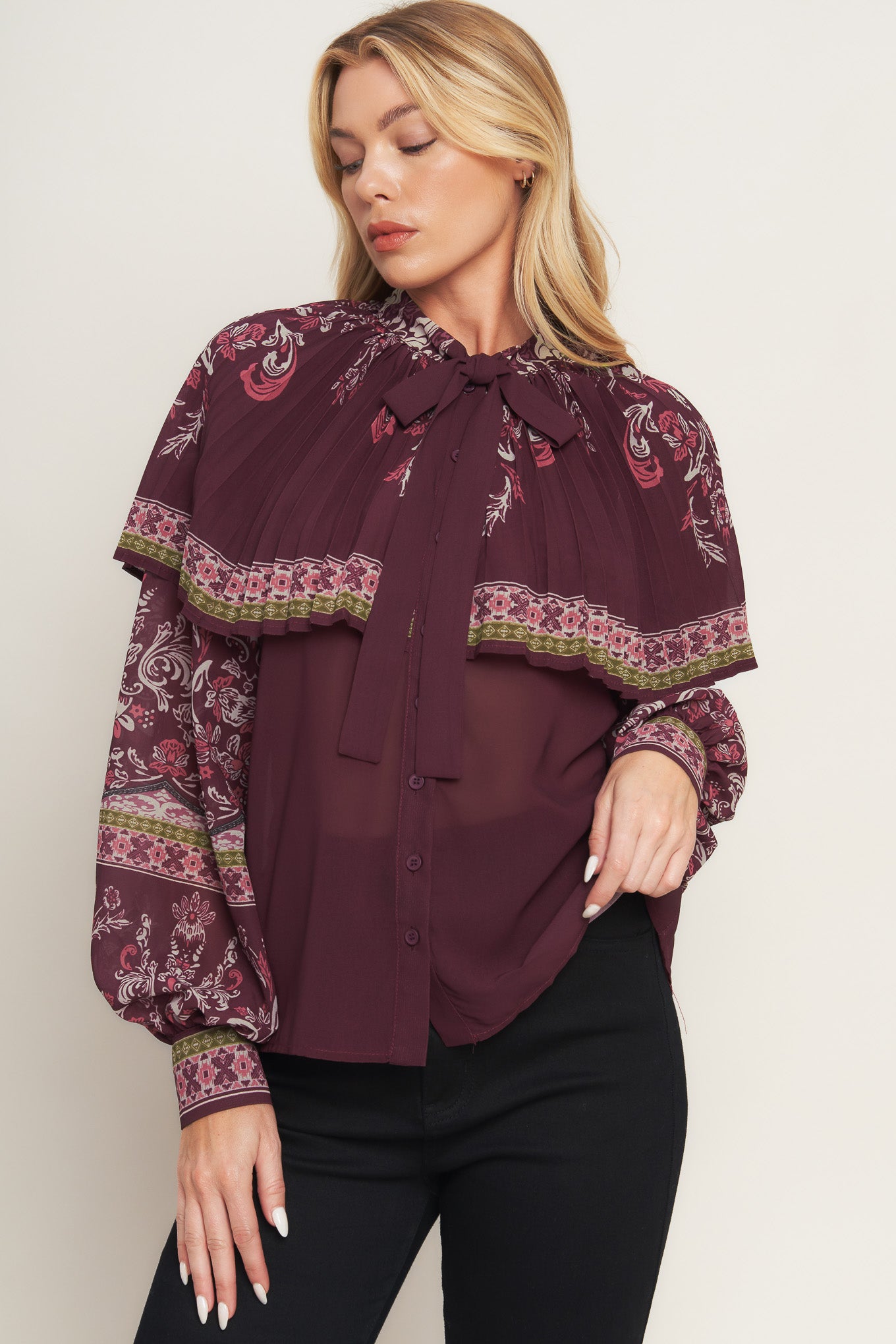 A printed woven top featuring pleated capelet, front bow tie, button down and long sleeve with cuff.