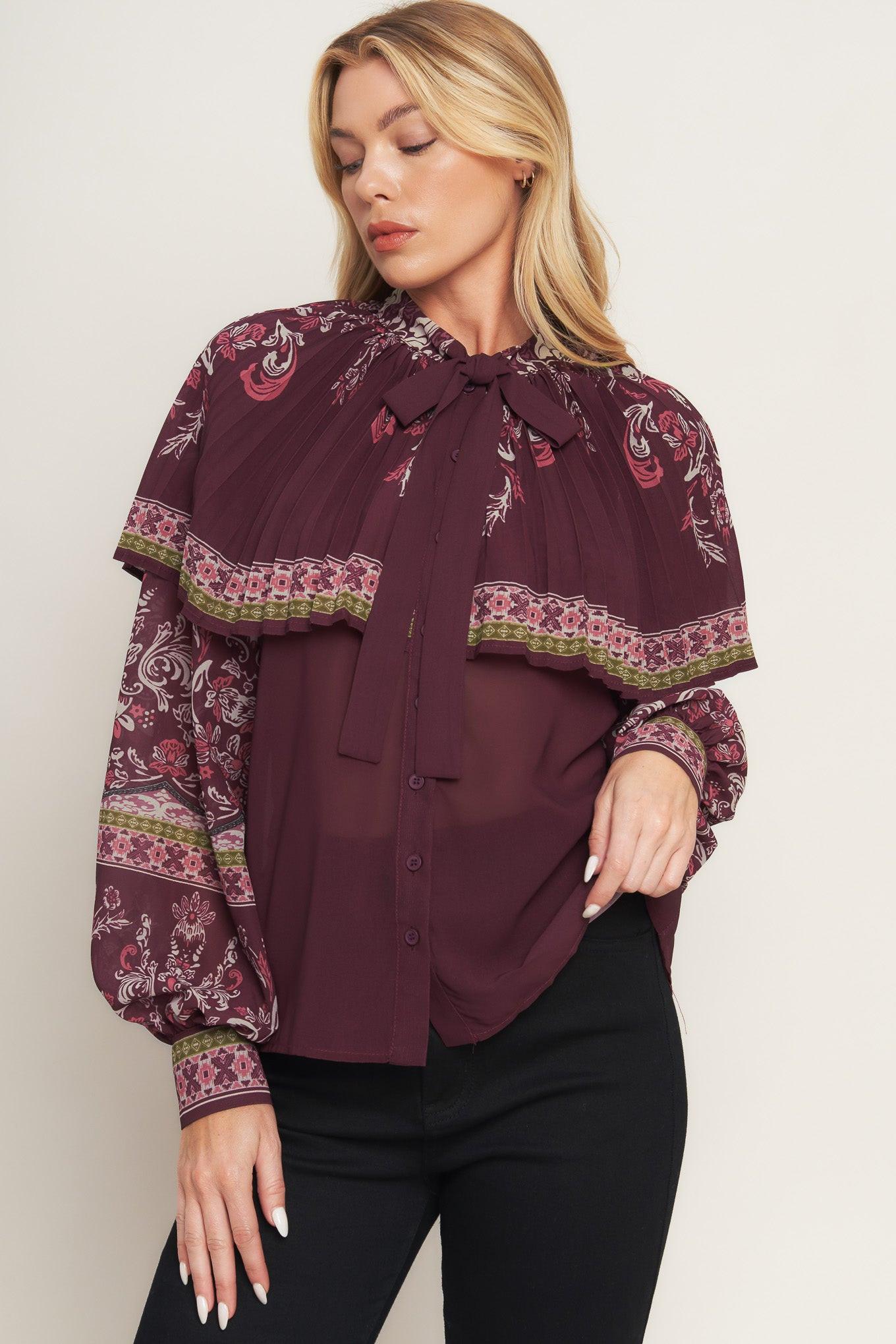 A printed woven top featuring pleated capelet, front bow tie, button down and long sleeve with cuff.