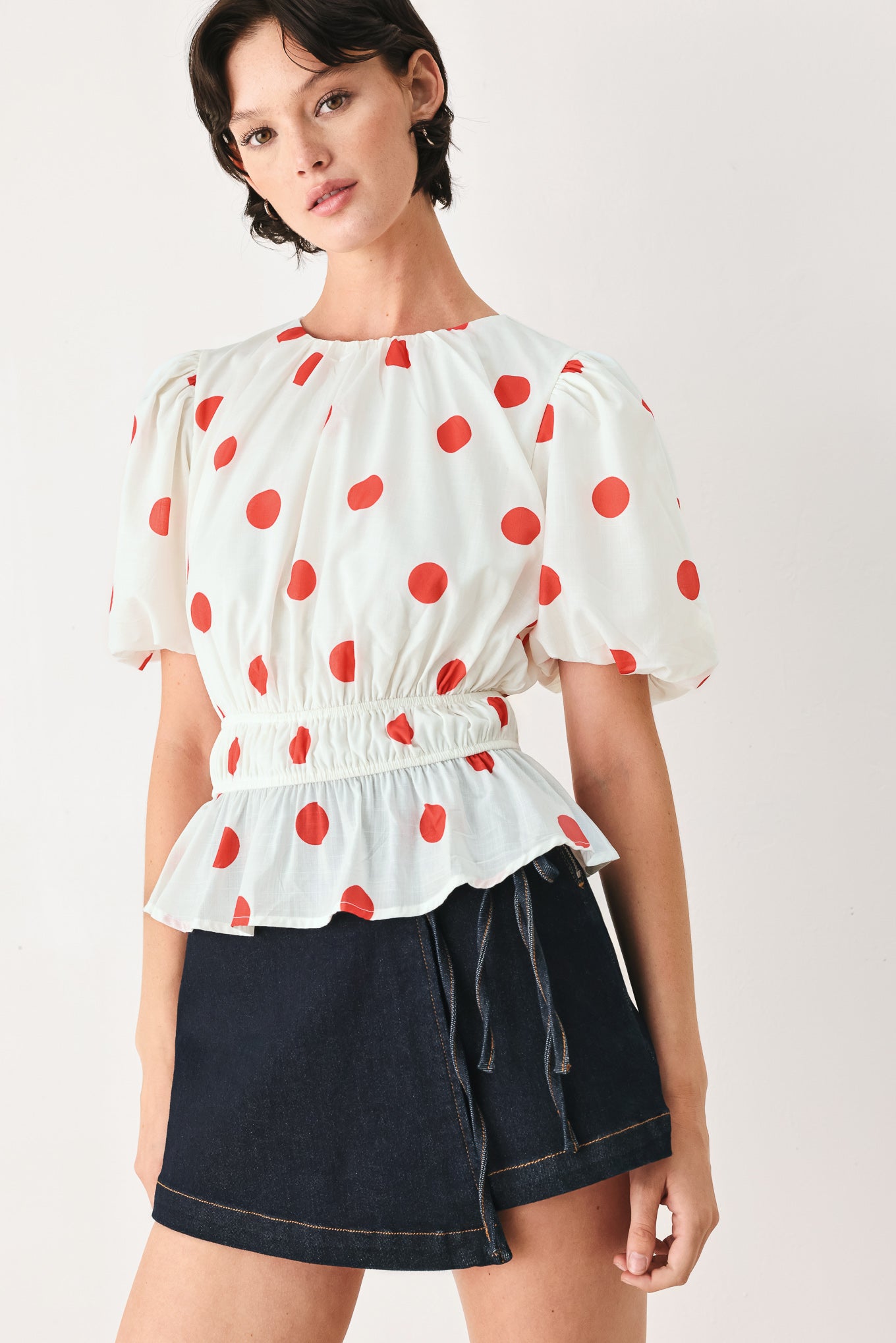 Woman wearing a white blouse with red polka dots featuring round neckline, short bubble sleeve, double elastic waist and back neck button closure | FREEMARKET