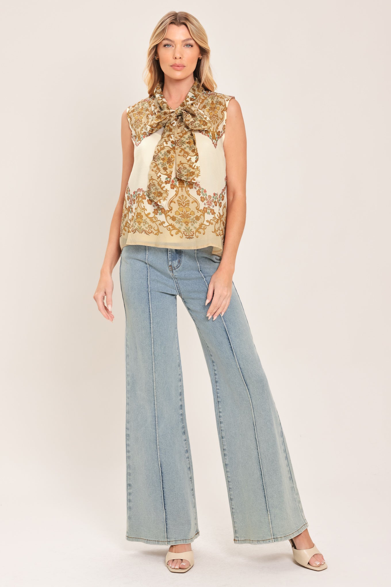 A woman wearing a sleeveless cream-colored blouse with a brown and gold damask pattern and a large bow tie-neck | Flying Tomato