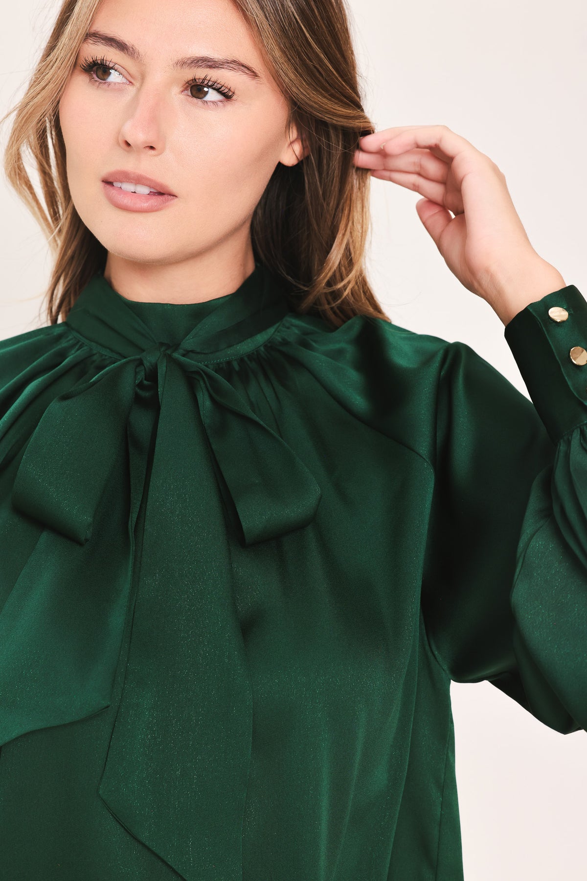 A woman wearing a dark hunter green satin blouse with a high neck, large bow at the collar, and gathered long sleeves with gold button accents | Flying Tomato