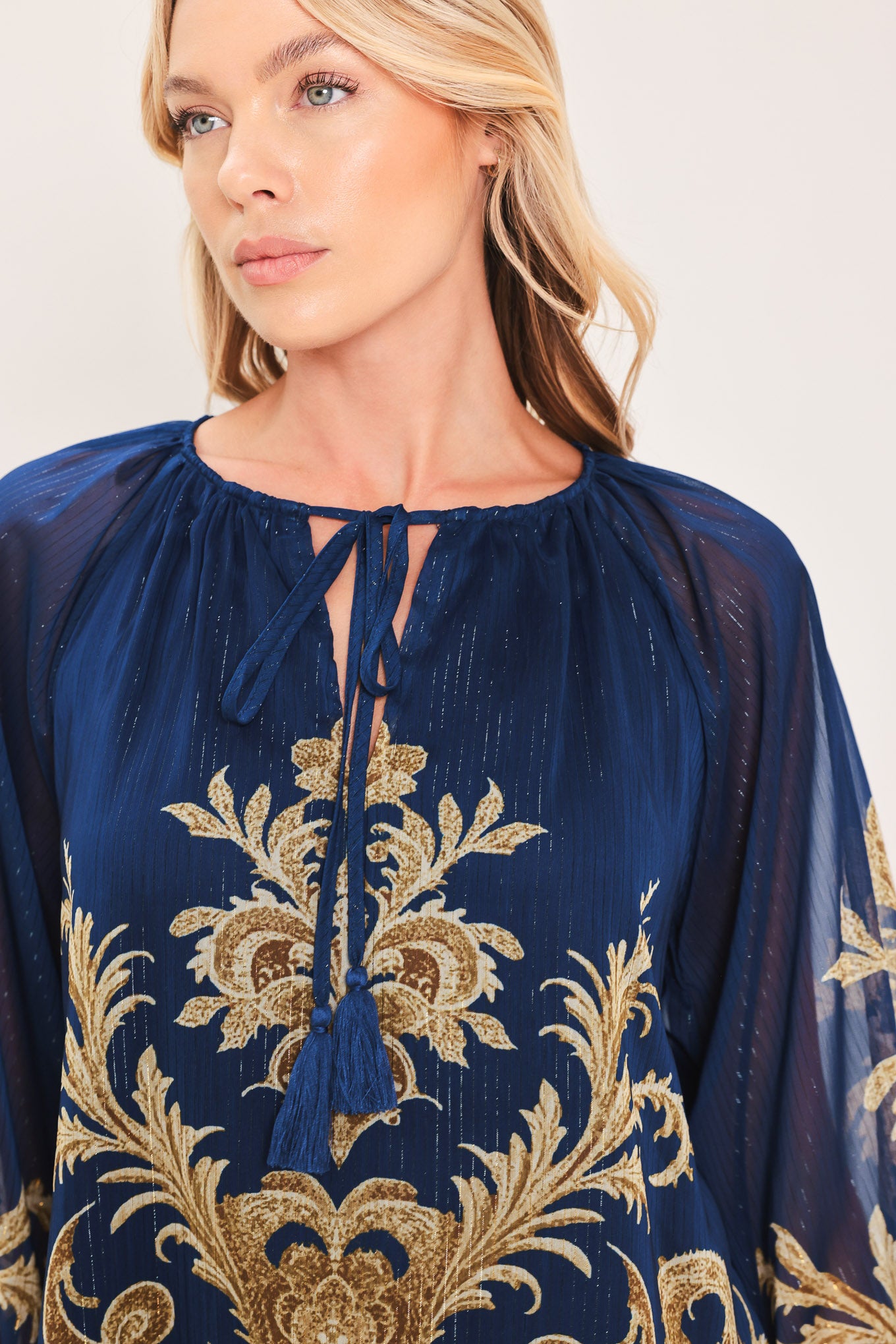 A woman wearing a navy blue long-sleeve blouse with an ornate gold baroque-style pattern and a tie-neck detail | Flying Tomato