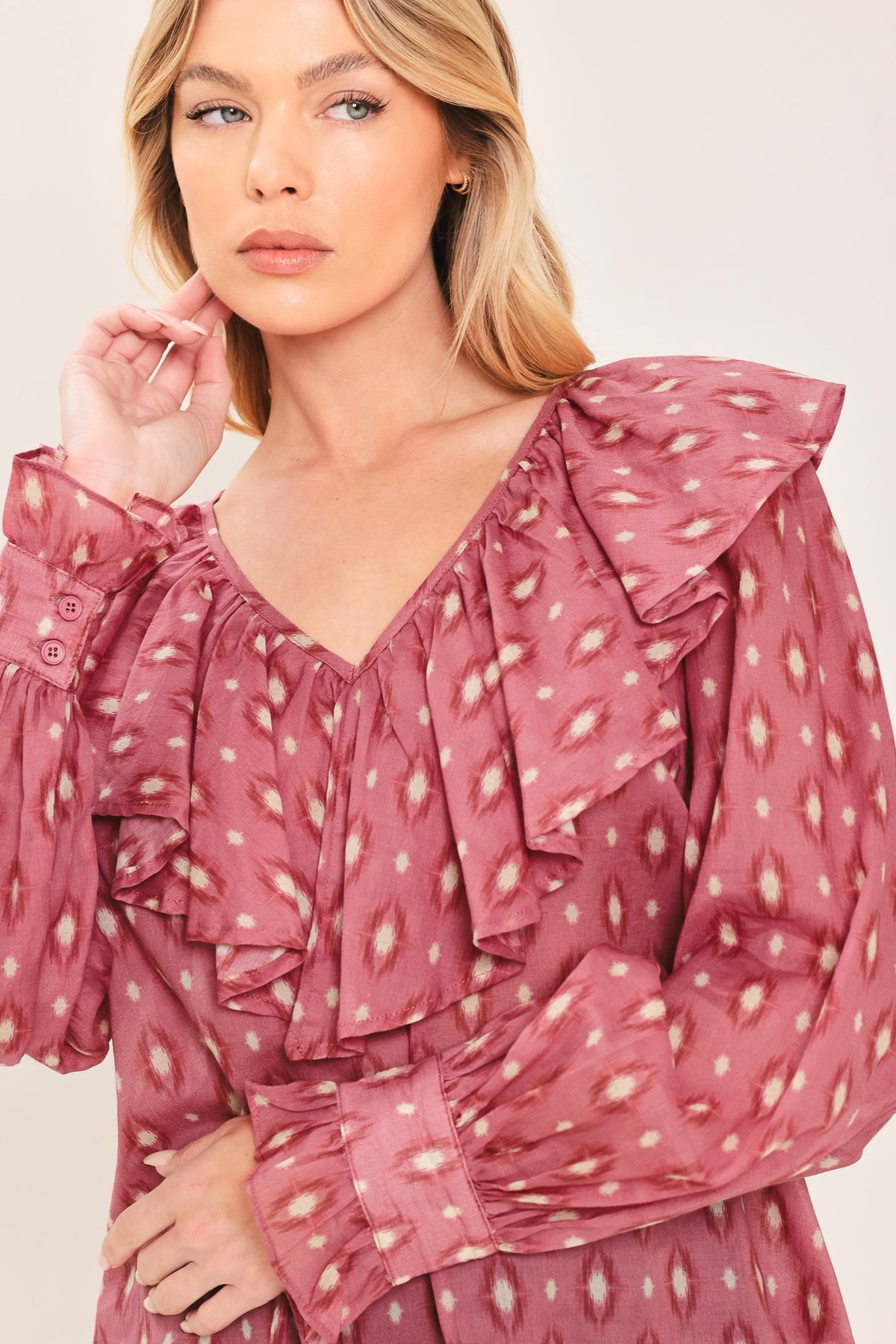 A woman wearing a dusty rose pink long-sleeve blouse with a white and beige ikat pattern, featuring layered ruffles around the V-neck and cuffs | Flying Tomato