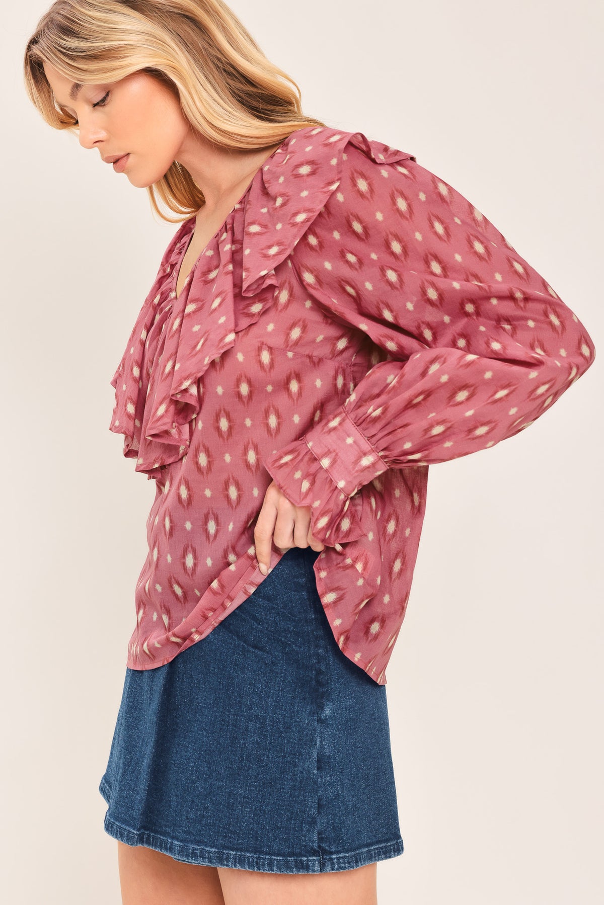 A woman wearing a dusty rose pink long-sleeve blouse with a white and beige ikat pattern, featuring layered ruffles around the V-neck and cuffs | Flying Tomato