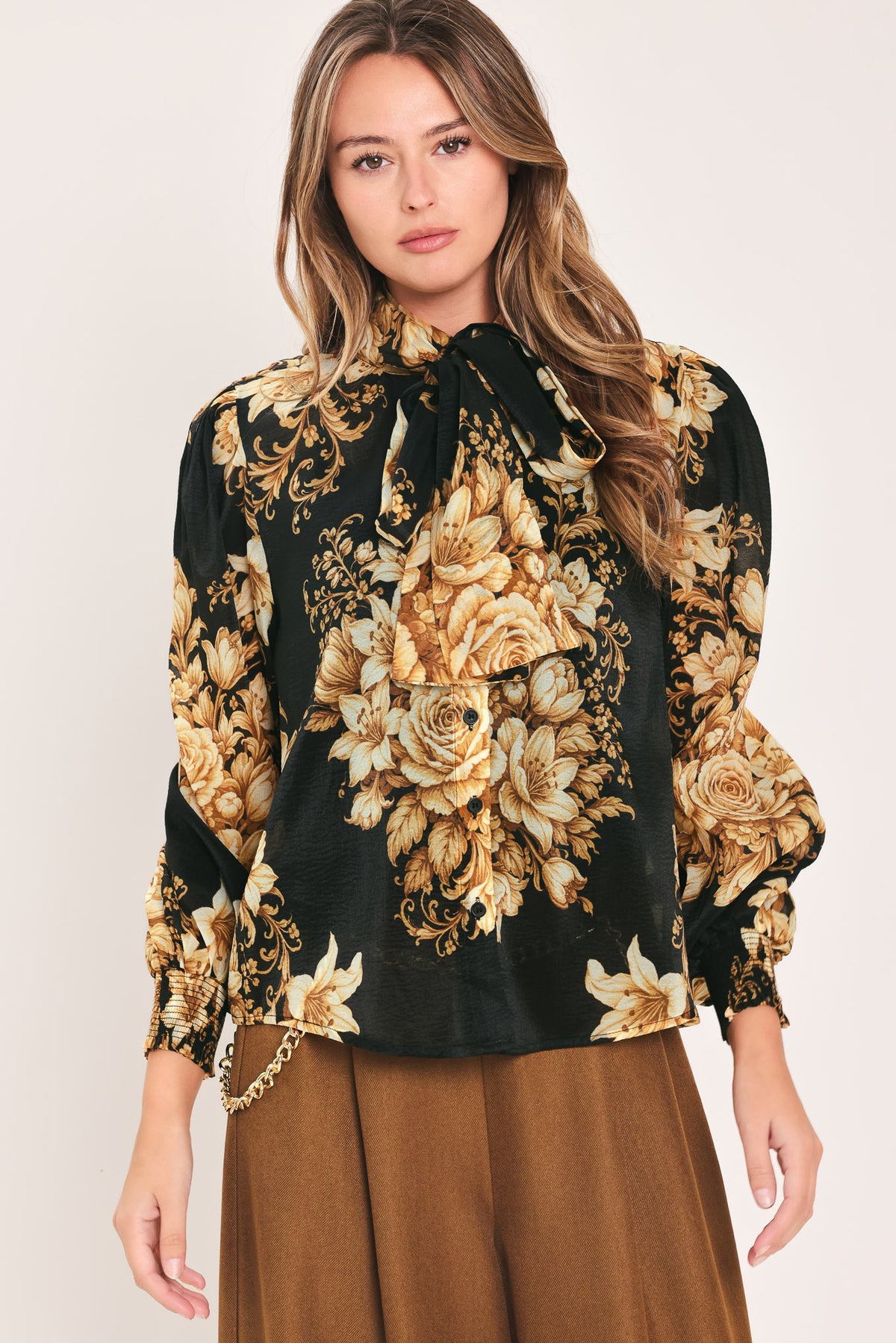 A woman wearing a black long-sleeve blouse with a large beige and gold floral print, featuring a high neck and bow detail | Flying Tomato