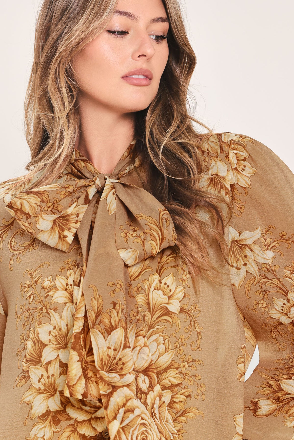 A woman wearing a sheer beige blouse with a large gold floral print and a bow tie neckline | Flying Tomato
