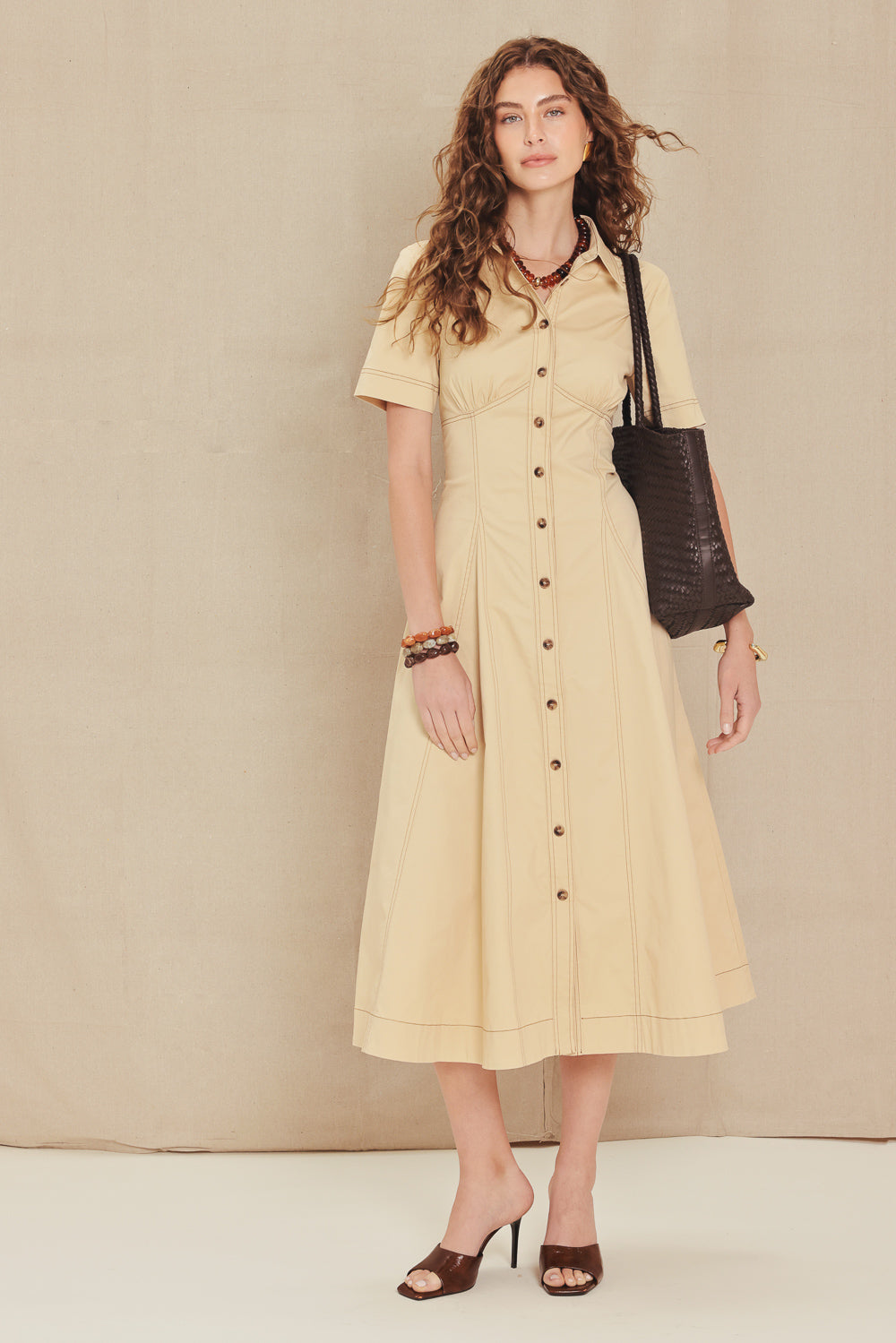 A woman models a short-sleeved cream-colored midi dress with a button-down front, standing against a neutral beige backdrop. She is carrying a dark brown textured shoulder bag and wearing brown heeled sandals.