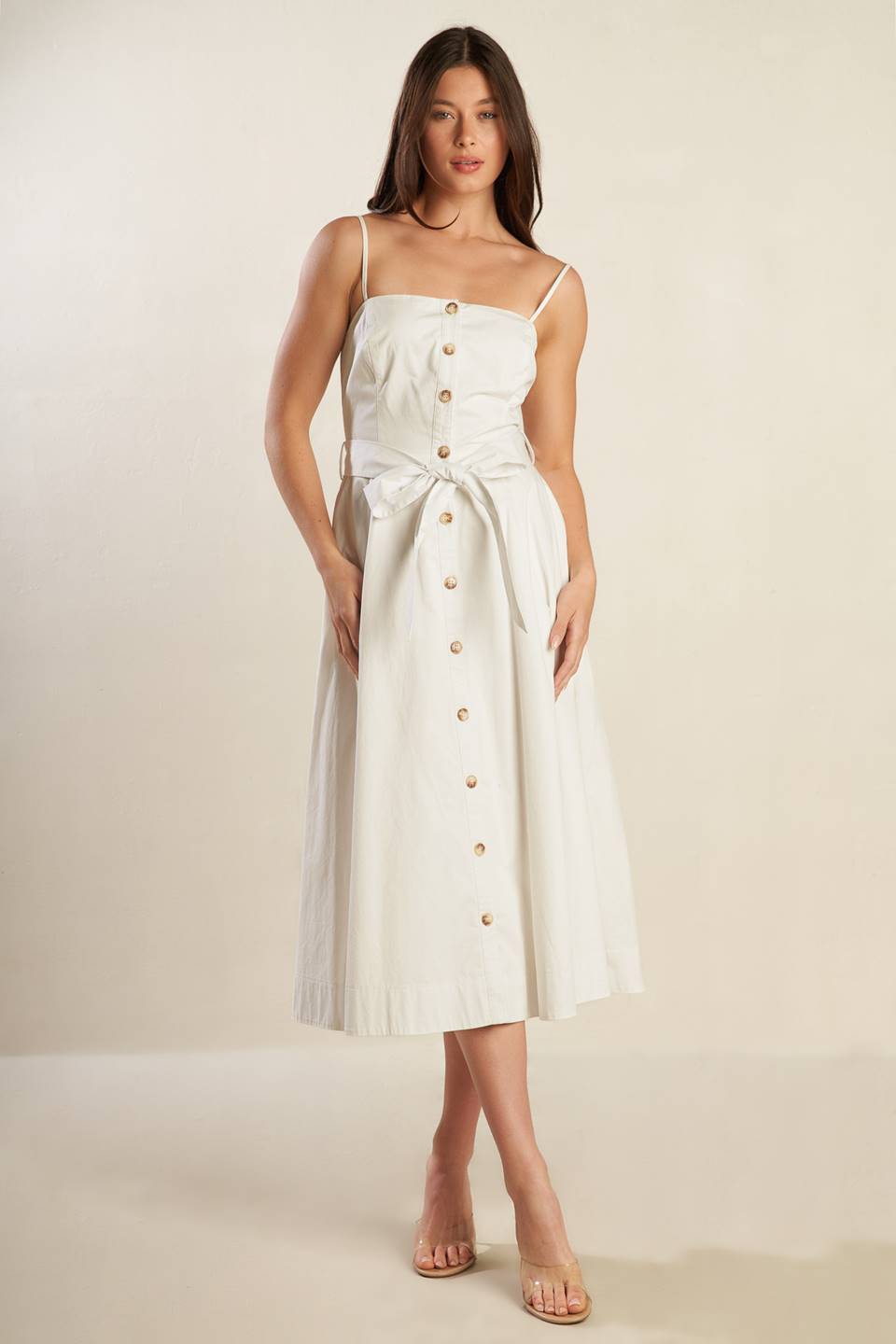 A solid white woven midi dress featuring straight neckline straps, button down, side pockets, self sash belt and full skirt.