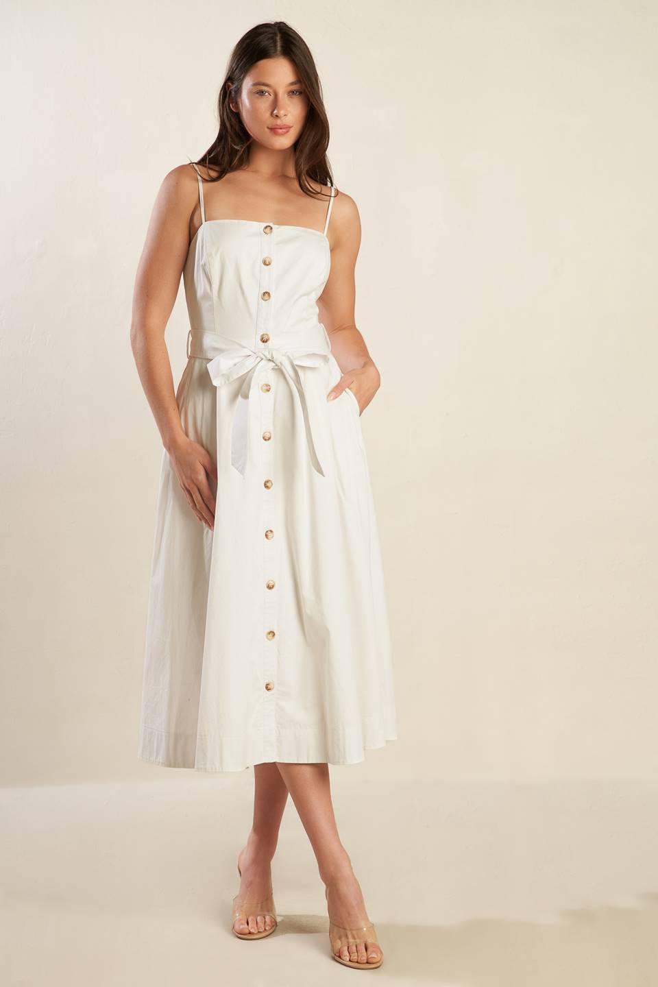 A solid white woven midi dress featuring straight neckline straps, button down, side pockets, self sash belt and full skirt.