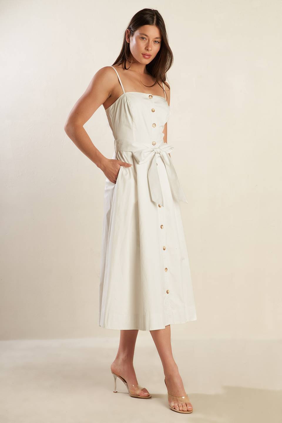 A solid white woven midi dress featuring straight neckline straps, button down, side pockets, self sash belt and full skirt.