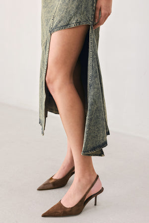 A woman modeling a sleeveless, mid-length denim dress with a distressed denim acid-wash finish, featuring a high side slit and brown pointed-toe heels | FREEMARKET