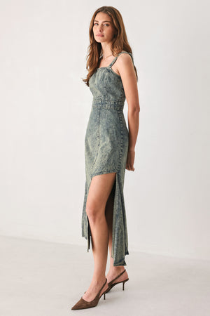 A woman modeling a sleeveless, mid-length denim dress with a distressed denim acid-wash finish, featuring a high side slit and brown pointed-toe heels | FREEMARKET