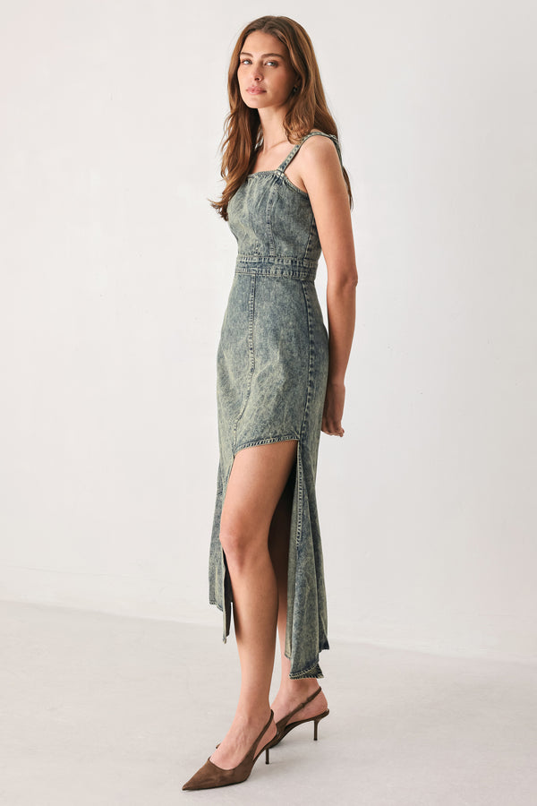 A woman modeling a sleeveless, mid-length denim dress with a distressed denim acid-wash finish, featuring a high side slit and brown pointed-toe heels | FREEMARKET