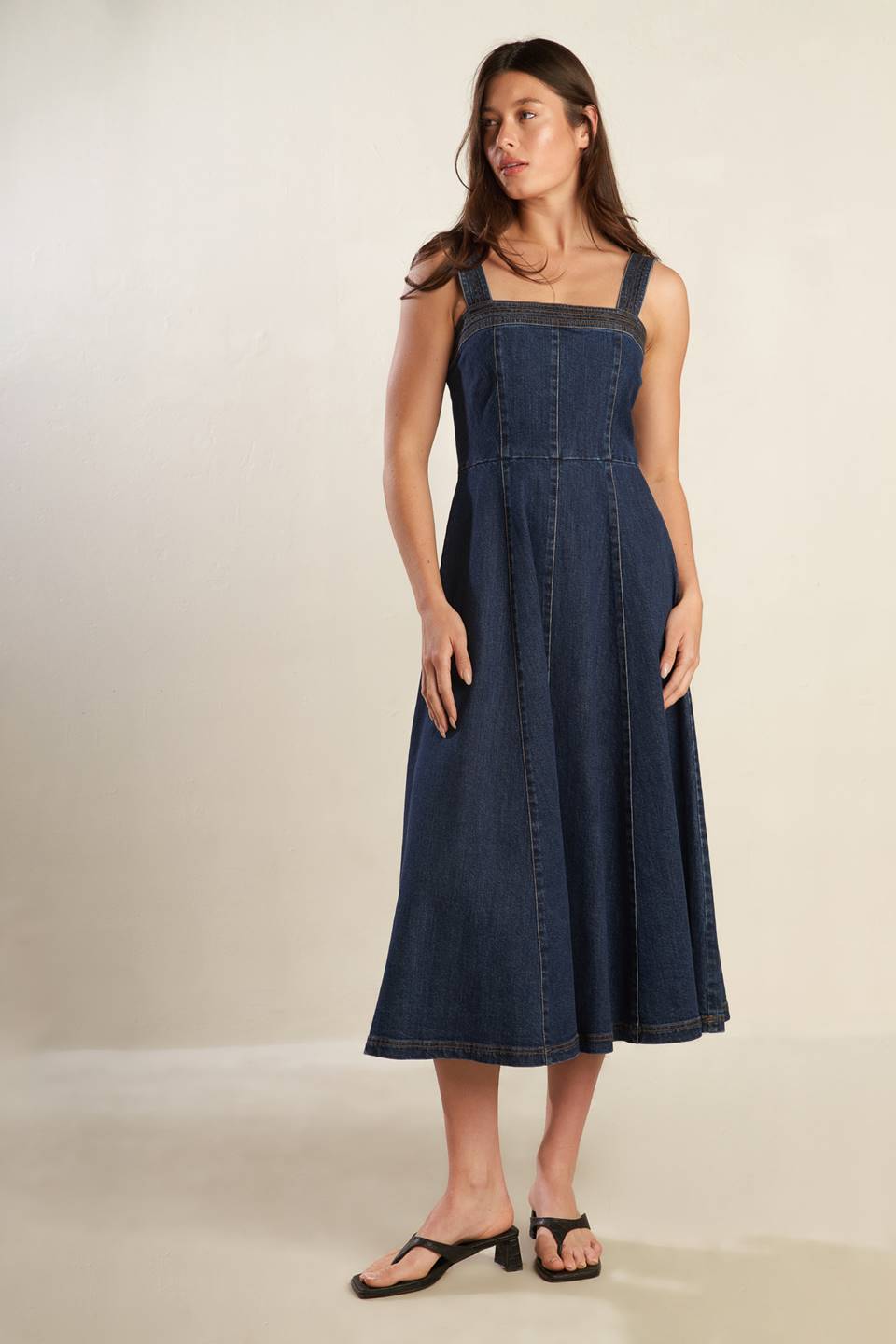 A dark washed denim midi dress featuring straight neckline, wide strap, full skirt and back zipper closure.