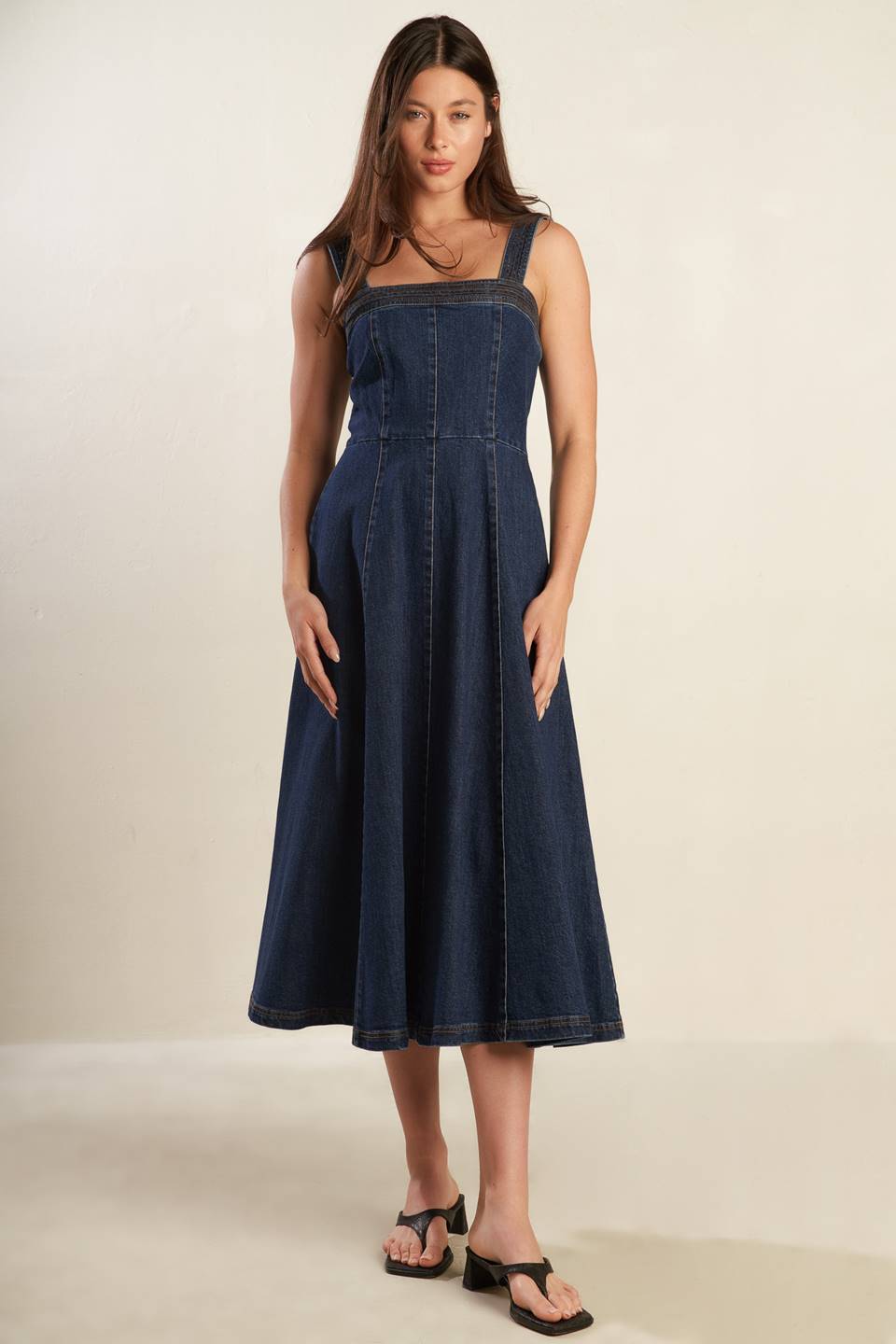 A dark washed denim midi dress featuring straight neckline, wide strap, full skirt and back zipper closure.