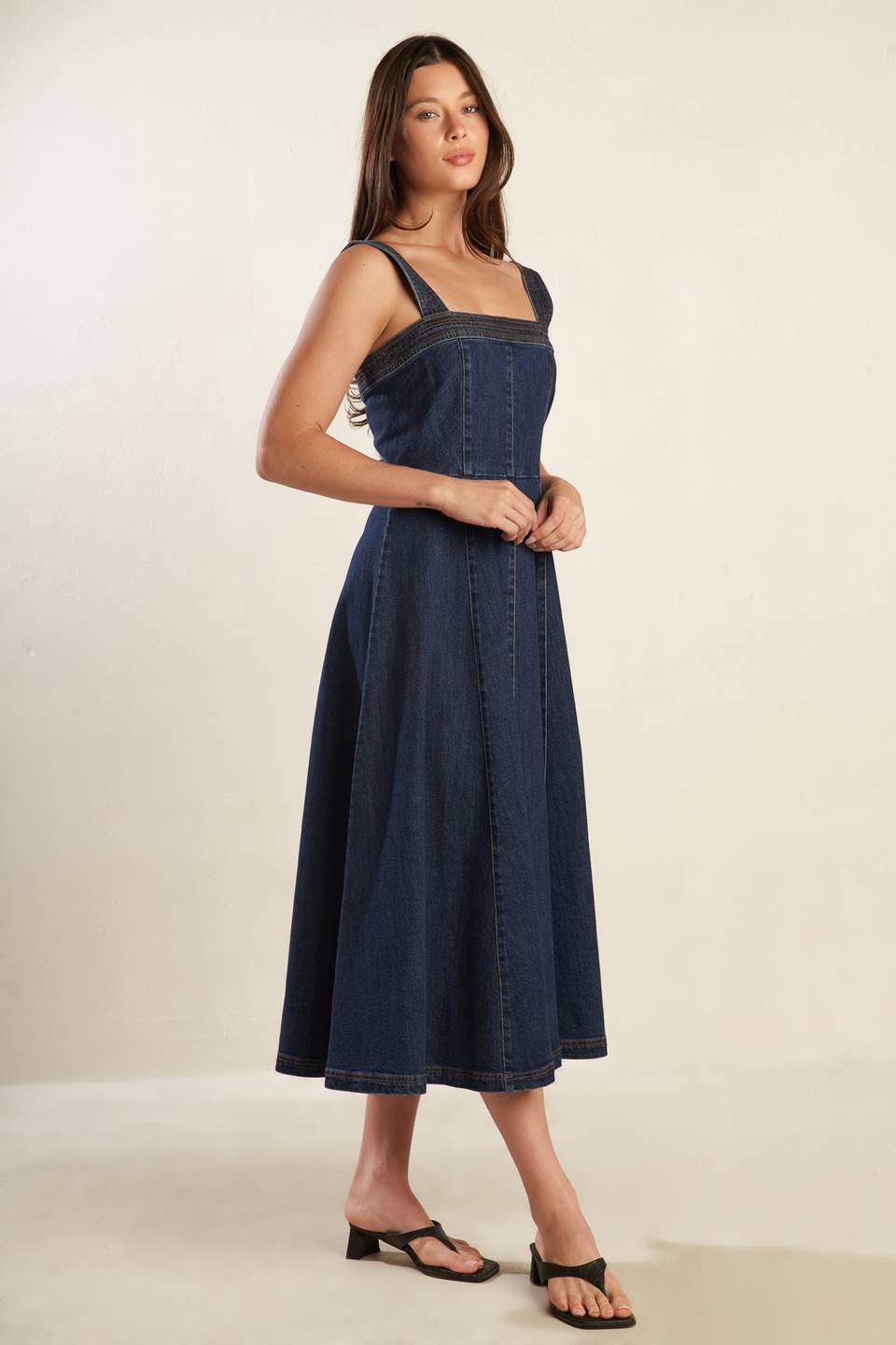 A dark washed denim midi dress featuring straight neckline, wide strap, full skirt and back zipper closure.