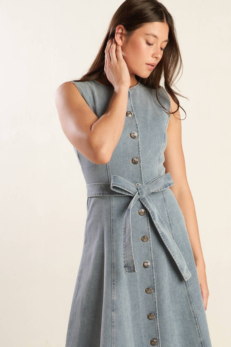 A sleeveless, washed light denim midi dress featuring round neckline, button down, self sash tie, side pockets and full skirt.