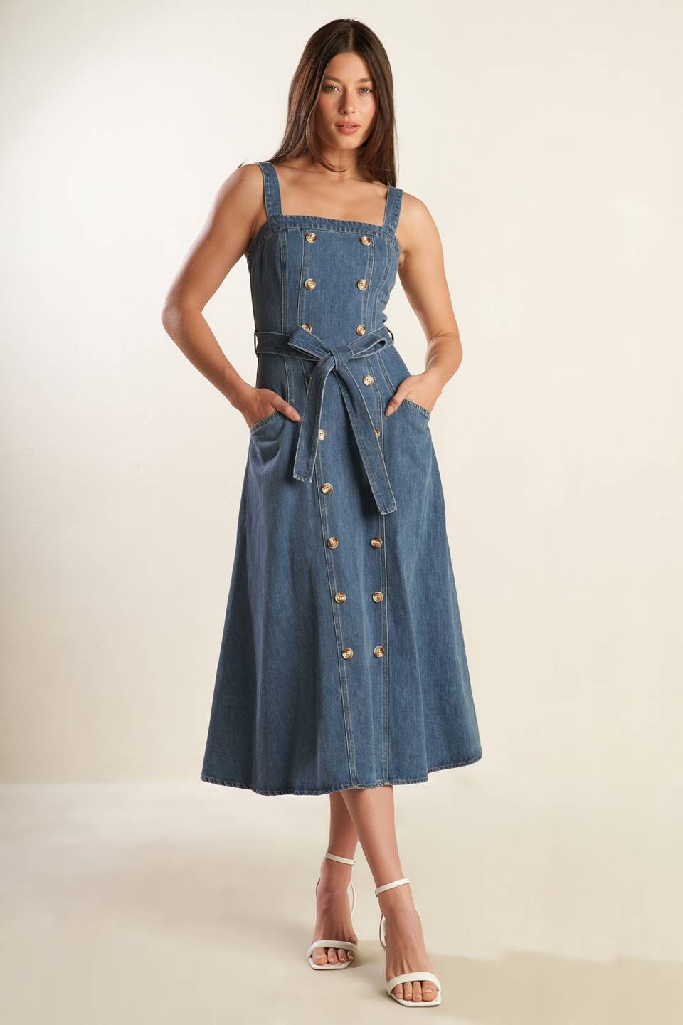 A washed blue denim midi dress featuring straight neckline, straps, double button down, self sash tie, side pockets and back zipper closure.