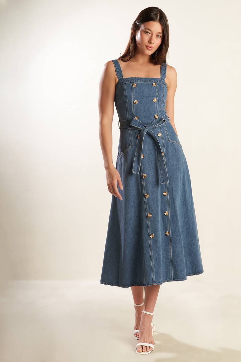 A washed blue denim midi dress featuring straight neckline, straps, double button down, self sash tie, side pockets and back zipper closure.