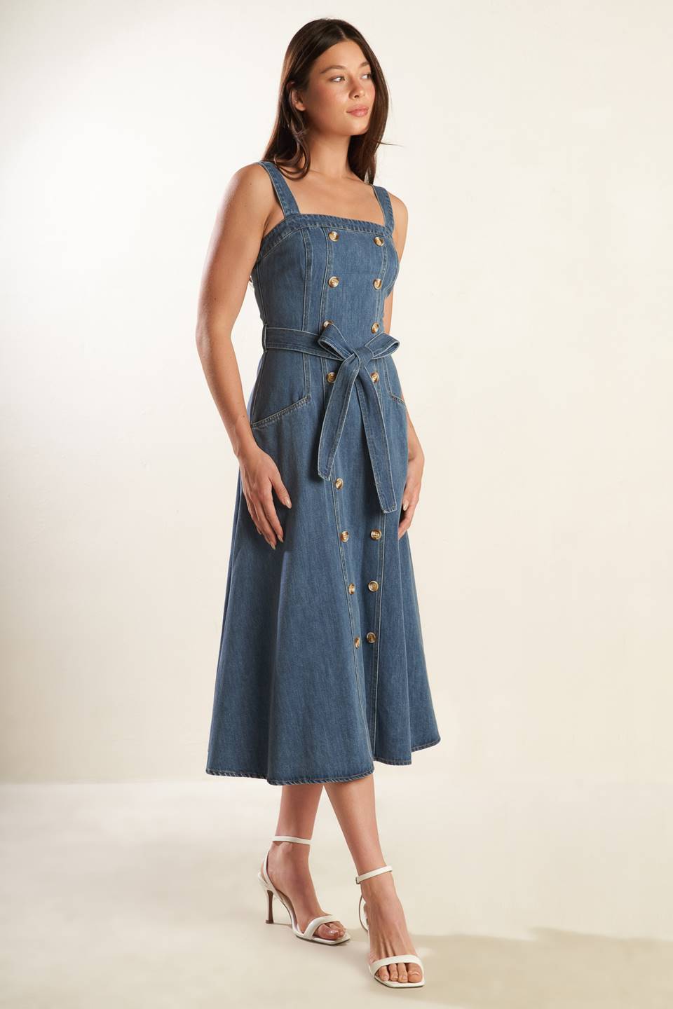 A washed blue denim midi dress featuring straight neckline, straps, double button down, self sash tie, side pockets and back zipper closure.