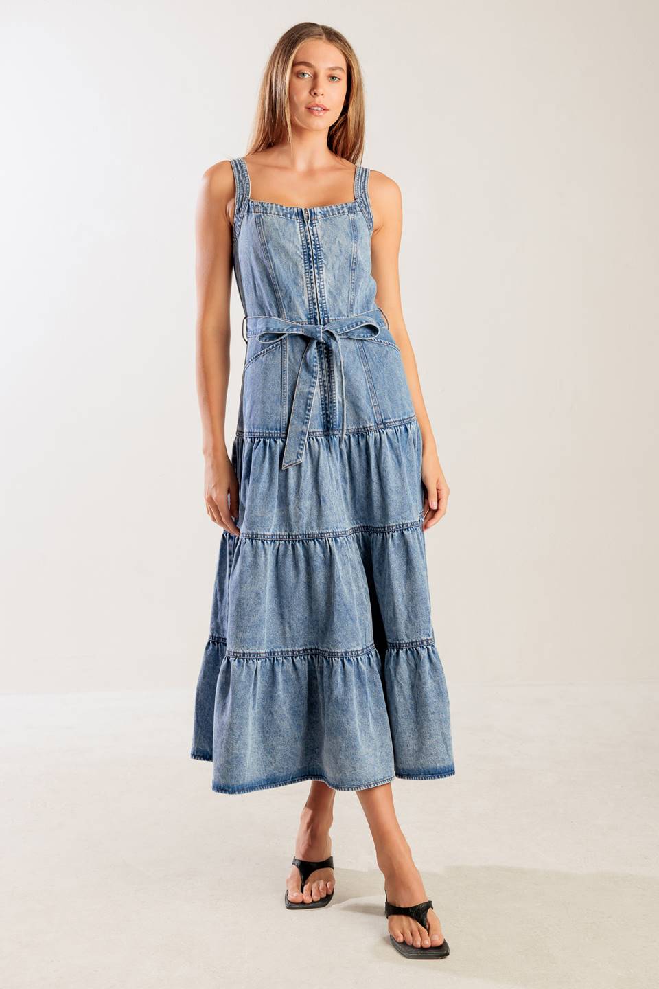 A washed denim dress featuring straight neckline, straps, front zipper closure, self sash tie and tiered skirt.