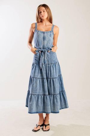 A washed denim dress featuring straight neckline, straps, front zipper closure, self sash tie and tiered skirt.