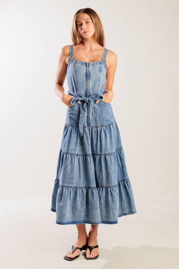A washed denim dress featuring straight neckline, straps, front zipper closure, self sash tie and tiered skirt.