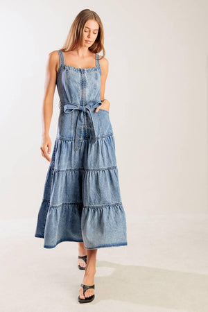 A washed denim dress featuring straight neckline, straps, front zipper closure, self sash tie and tiered skirt.