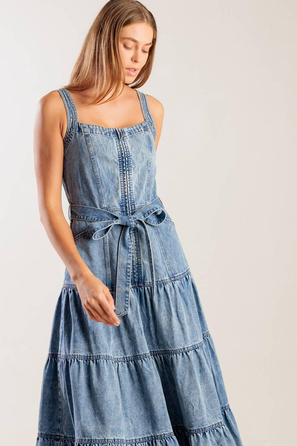 A washed denim dress featuring straight neckline, straps, front zipper closure, self sash tie and tiered skirt.
