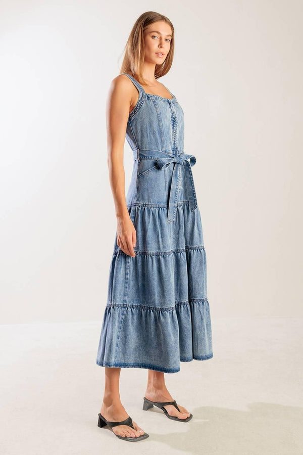 A washed denim dress featuring straight neckline, straps, front zipper closure, self sash tie and tiered skirt.