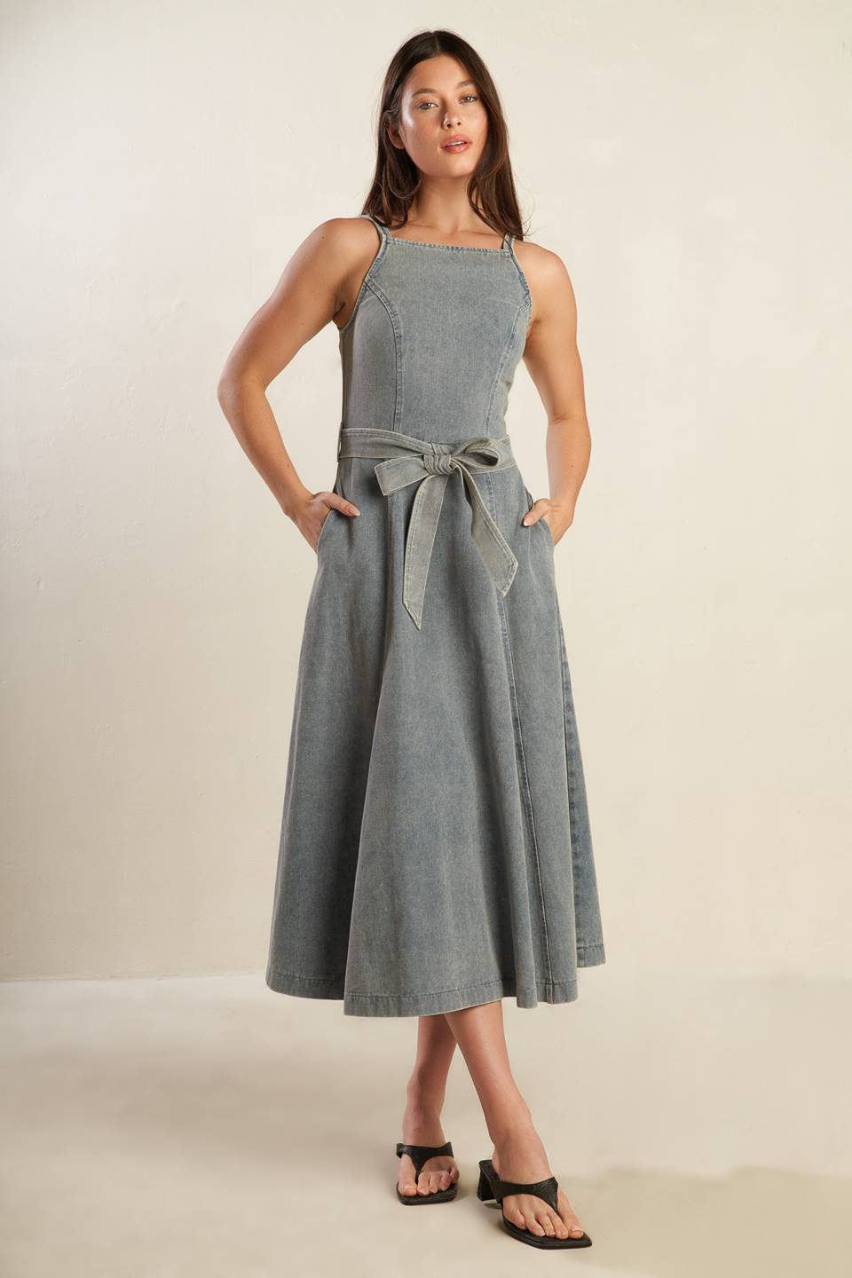 A washed denim midi dress featuring straight neckline, straps, circle skirt, self sash tie and back zipper closure.