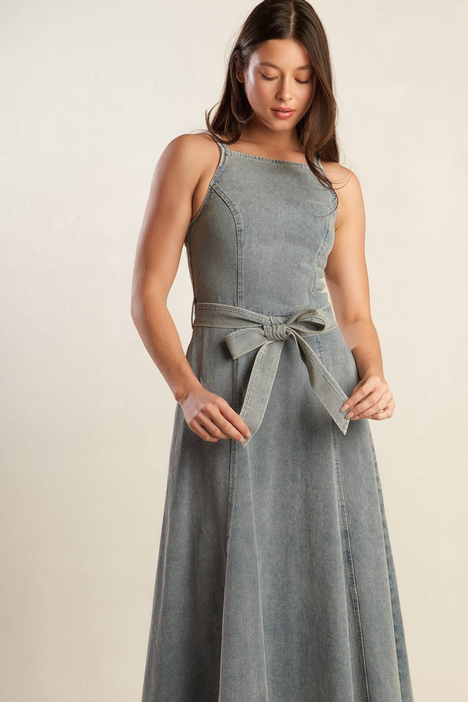 A washed denim midi dress featuring straight neckline, straps, circle skirt, self sash tie and back zipper closure.